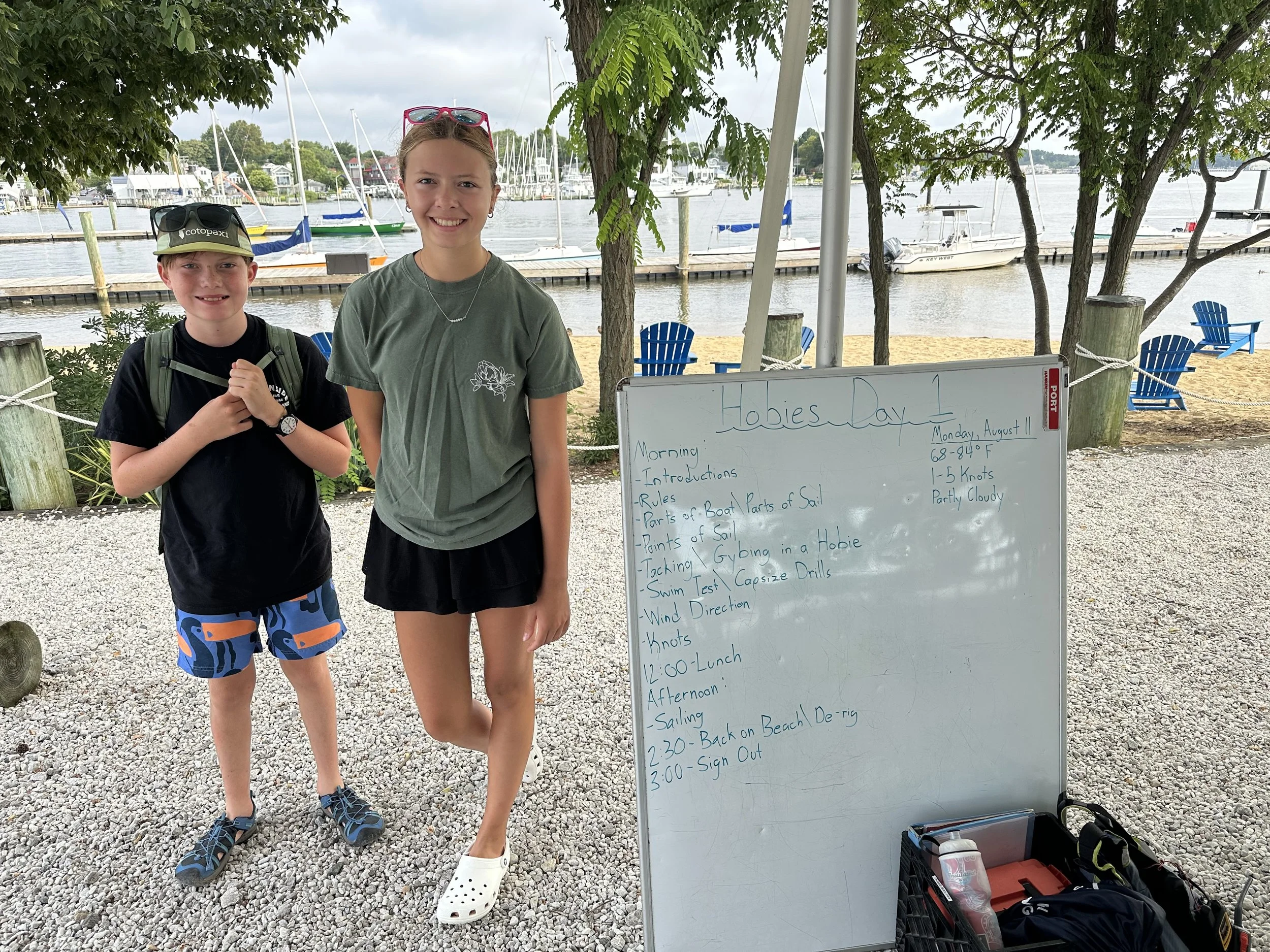Two children standing outdoors near a lake with boats docked in the background. A whiteboard with a schedule for 'Hobie Day' is visible next to them, and a black crate with equipment is in the foreground. Both children are smiling, wearing casual summer clothing.
