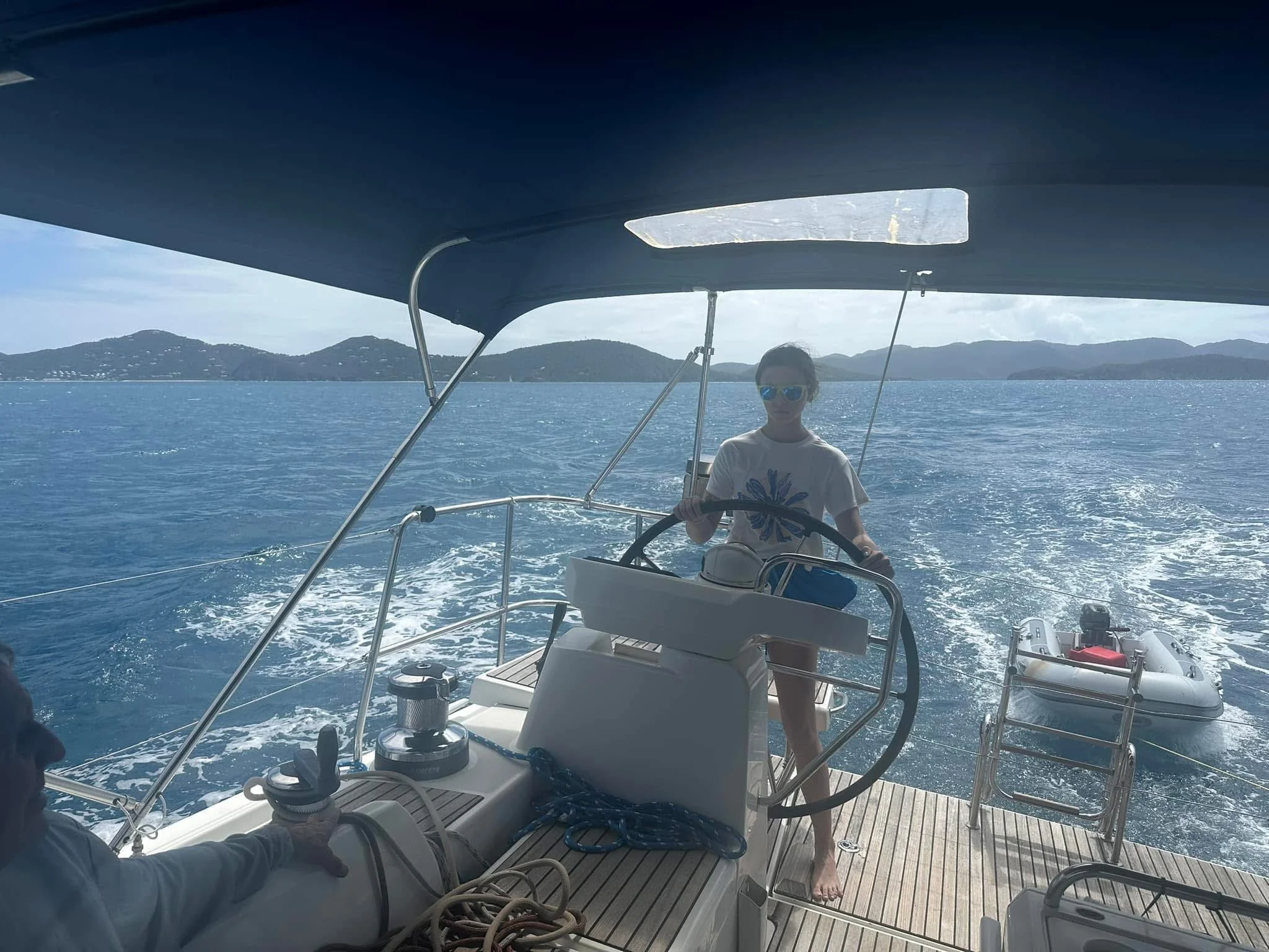 A woman steering a sailboat on the water, with a small motorized dinghy in the background, and a scenic landscape of hills and ocean in the distance.