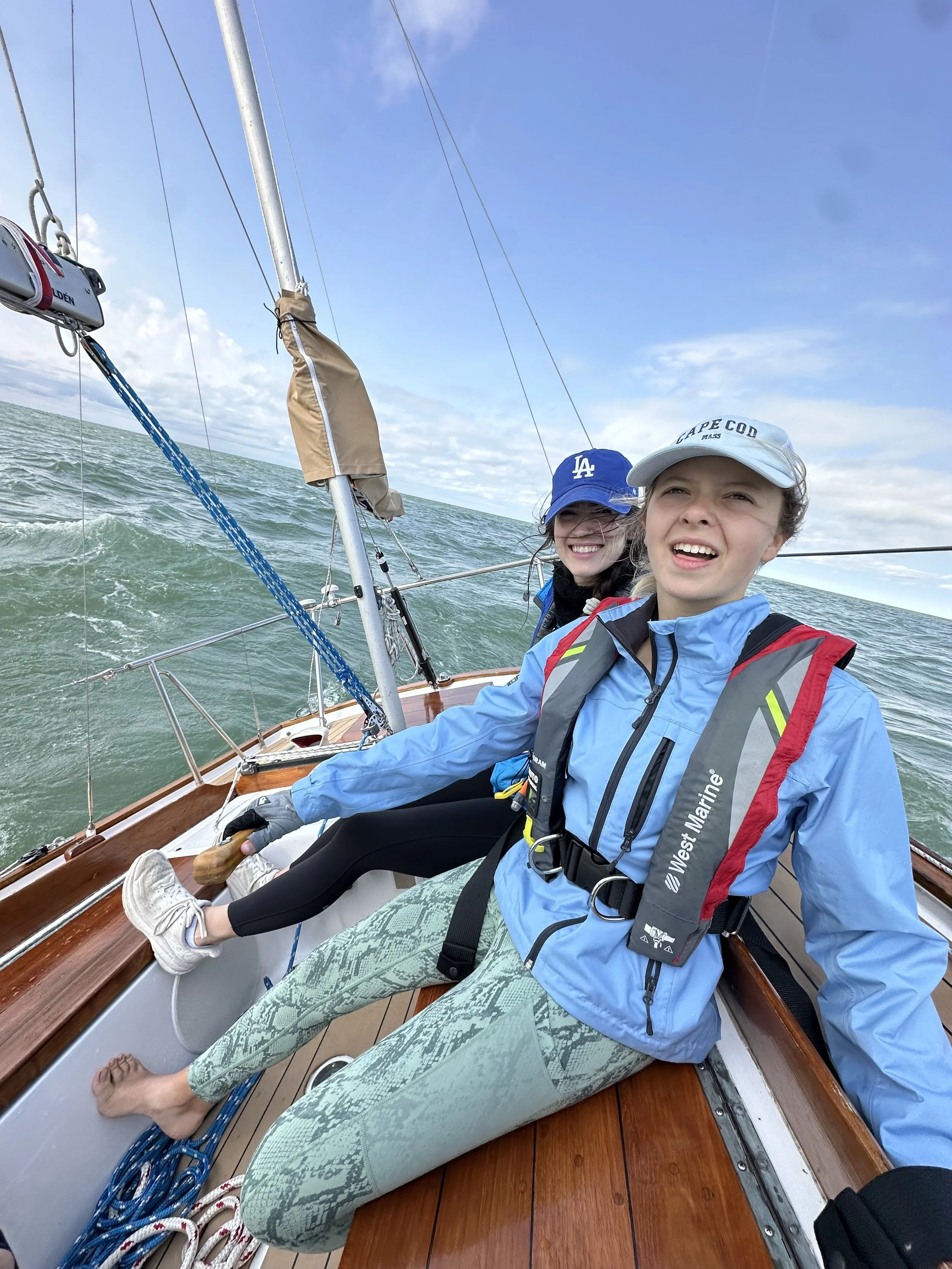 Two women sailing on a boat in the ocean, smiling and enjoying the day, wearing sailing gear and casual clothes.