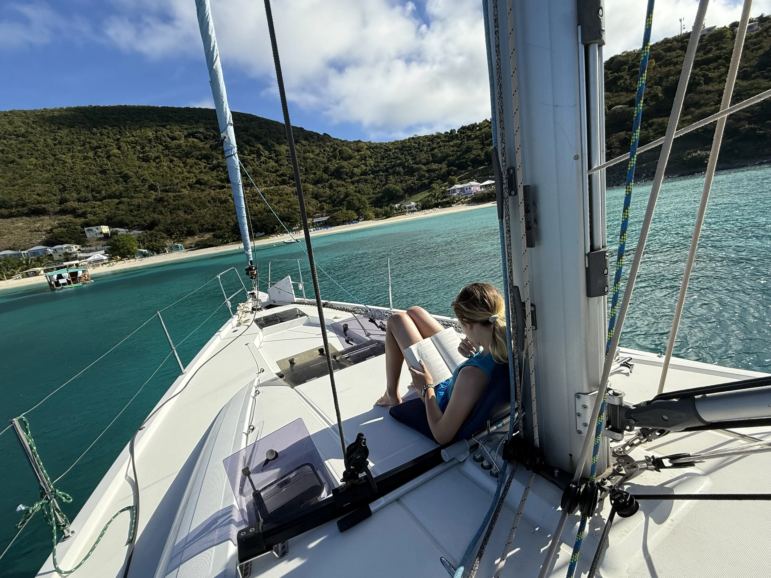 Woman reading a book while lounging on the deck of a sailboat near a tropical shoreline with hills, houses, and turquoise water.