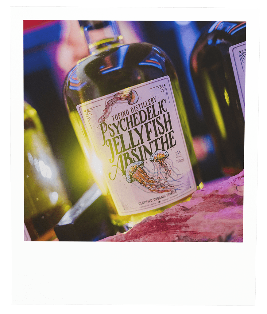 A bottle of Psychedelic Jellyfish Absinthe from Tofino Distillery with a colorful label featuring jellyfish illustrations, placed on a pink surface with other bottles in the background.