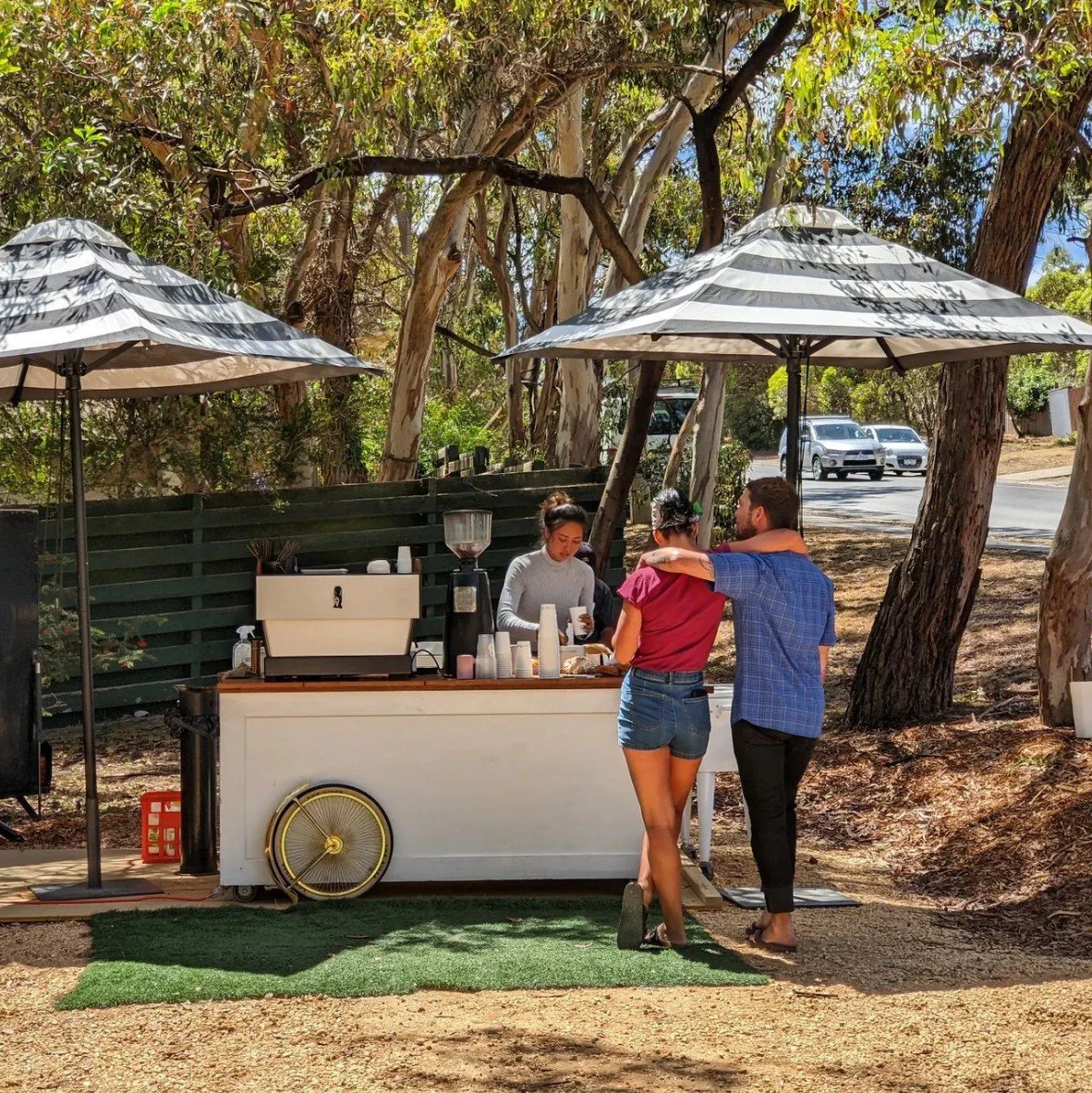 People ordering coffee at an outdoor coffee stand shaded by large umbrellas, with trees in the background and parked cars on the street.