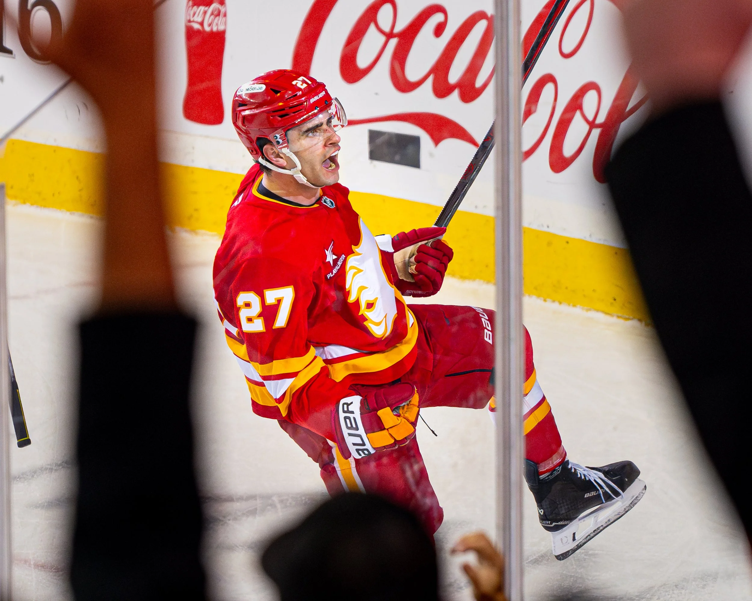 An ice hockey player wearing a red jersey with the number 27, red helmet, and holding a hockey stick, appears to be celebrating after a play, with his mouth open in a shout, on the ice near the boards with Coca-Cola signage.
