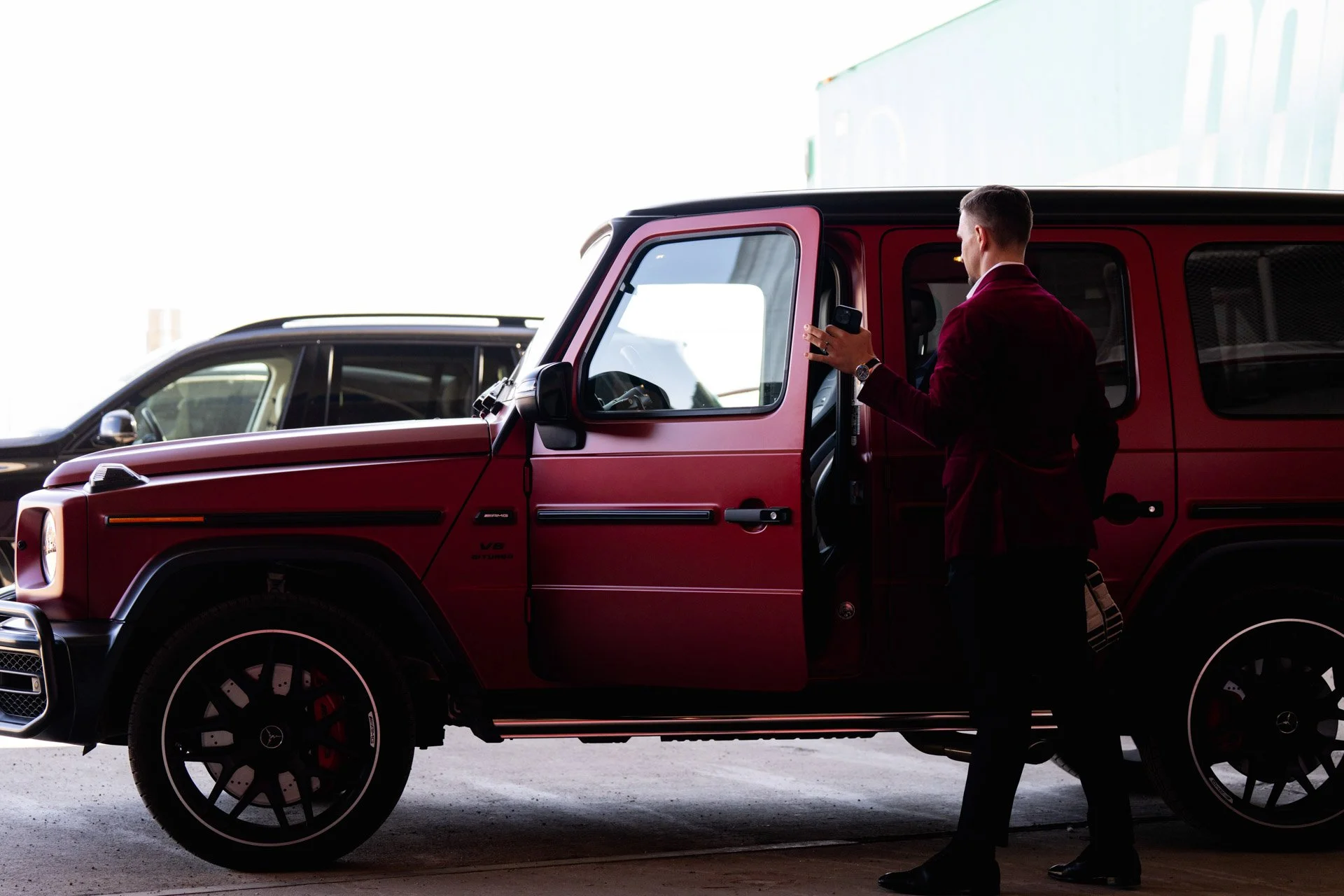 A man in a red jacket stands next to a red luxury SUV, looking at his phone.