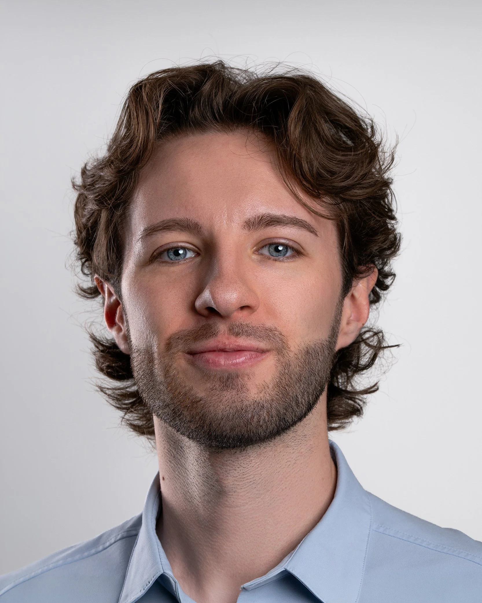 Close-up of a young man with wavy brown hair, blue eyes, and light skin, wearing a light blue collared shirt, against a plain grey background.