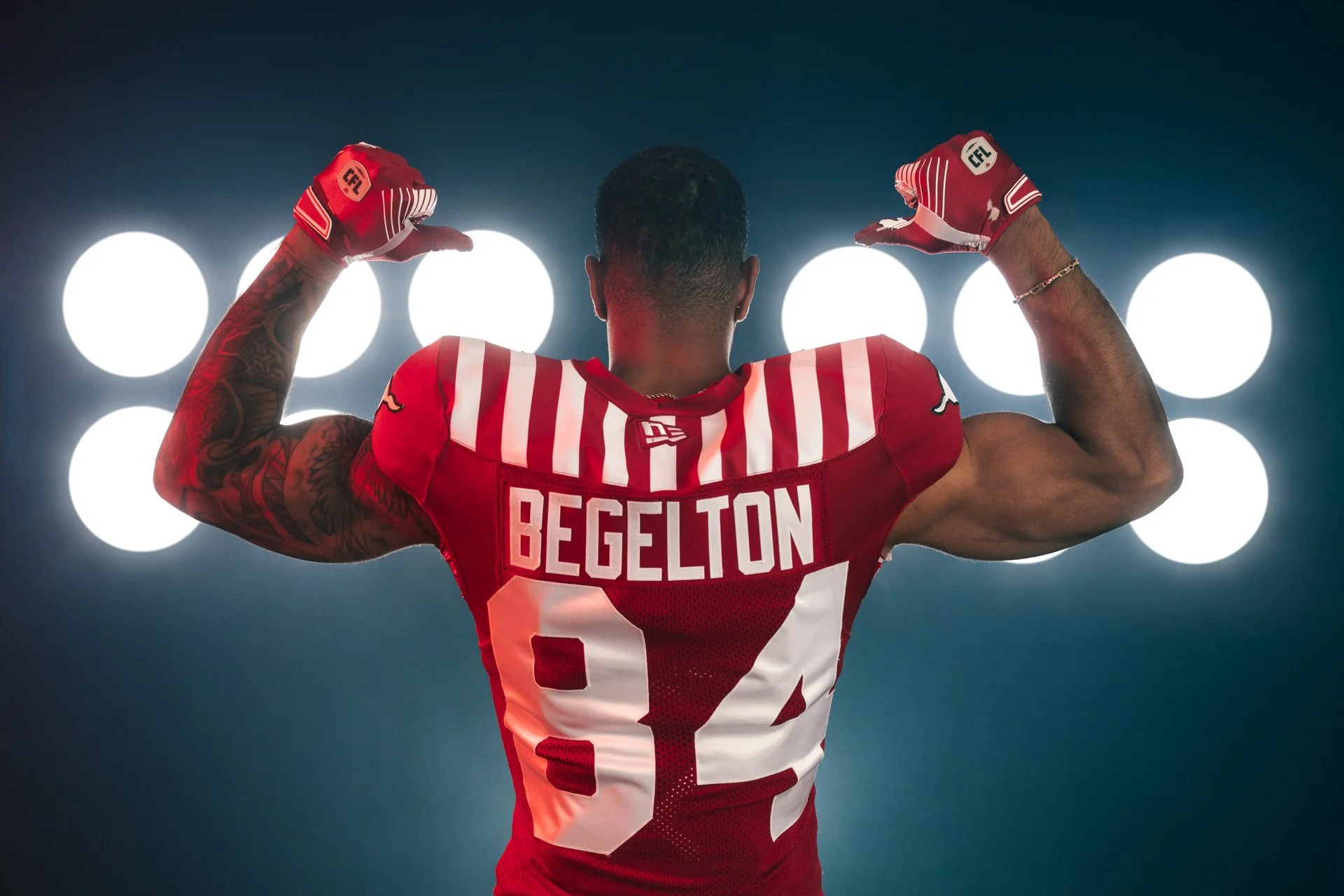 An American football player, wearing a red and white jersey with the name Begelton and the number 84, is flexing his biceps in front of bright circular lights.