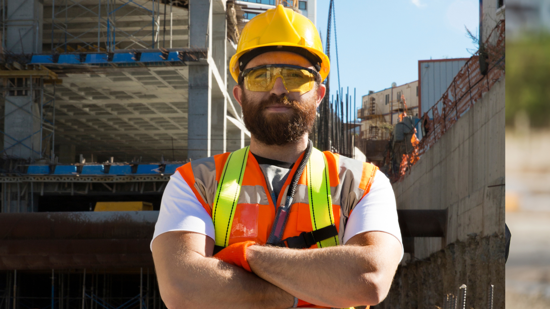 Construction worker with a beard, wearing a yellow hard hat, yellow safety glasses, and an orange safety vest, standing with arms crossed at a construction site background.