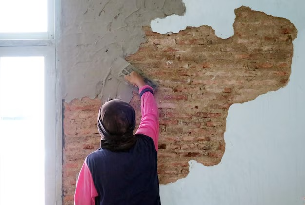 A person wearing black gloves and white sneakers is installing wooden floor tiles using a mallet. The person is kneeling on a concrete floor, and entire floor tiles are being aligned with small yellow spacers. A large bucket of adhesive or grout and a trowel are visible nearby.