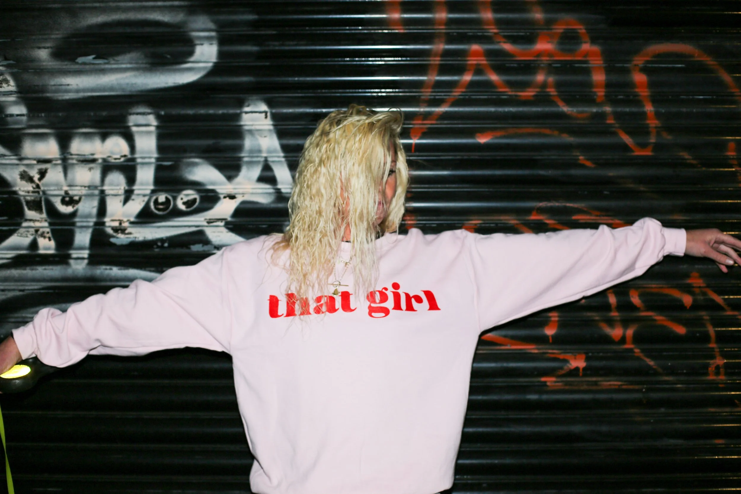 A woman with blonde, curly hair wearing a white sweatshirt with red text that reads 'that girl,' standing with arms outstretched in front of a black corrugated metal wall with graffiti.