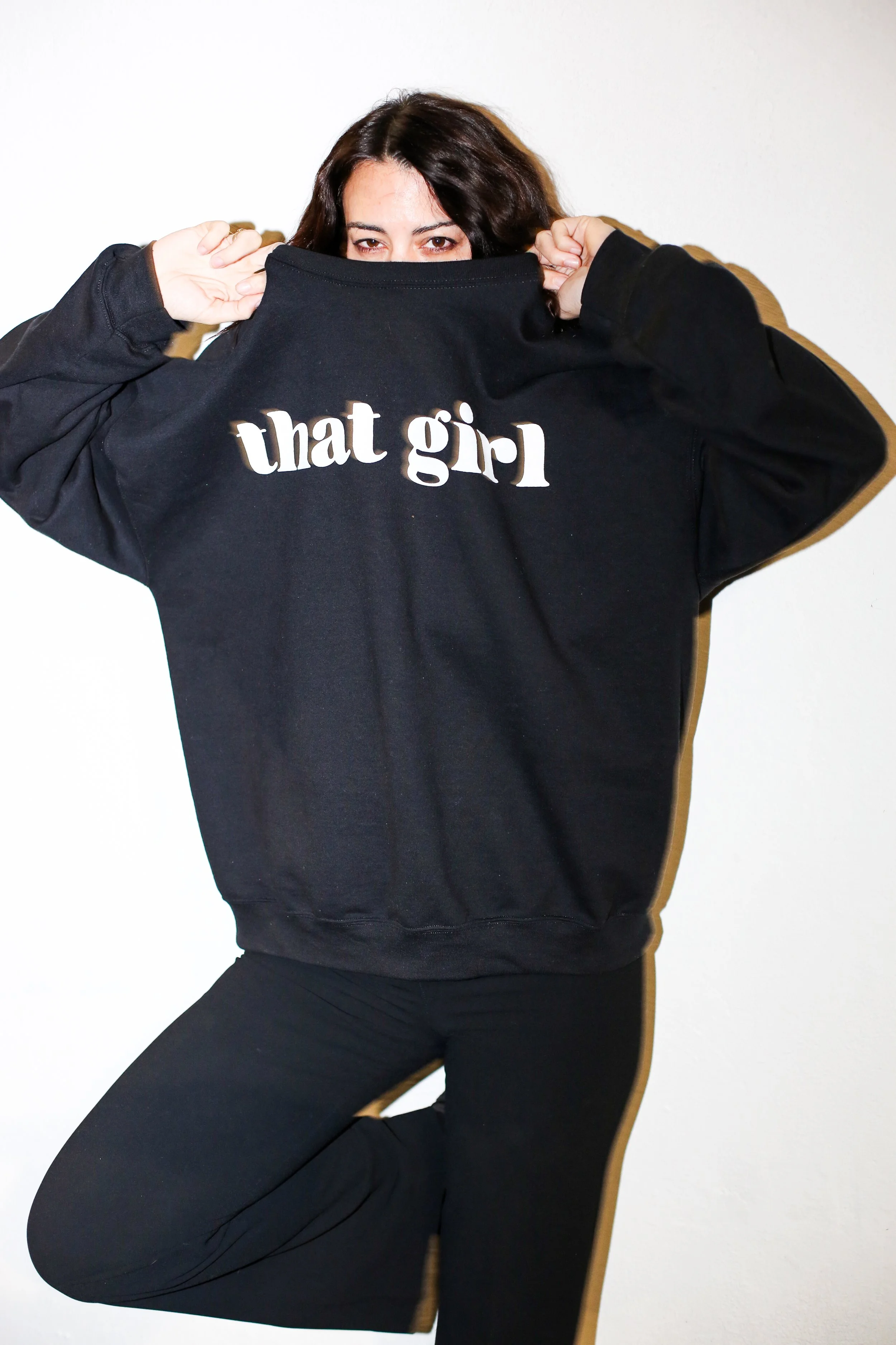 Person with dark hair wearing a black hoodie with the words 'that girl' on the back, partially lifting the hoodie to cover their face, standing against a white background.