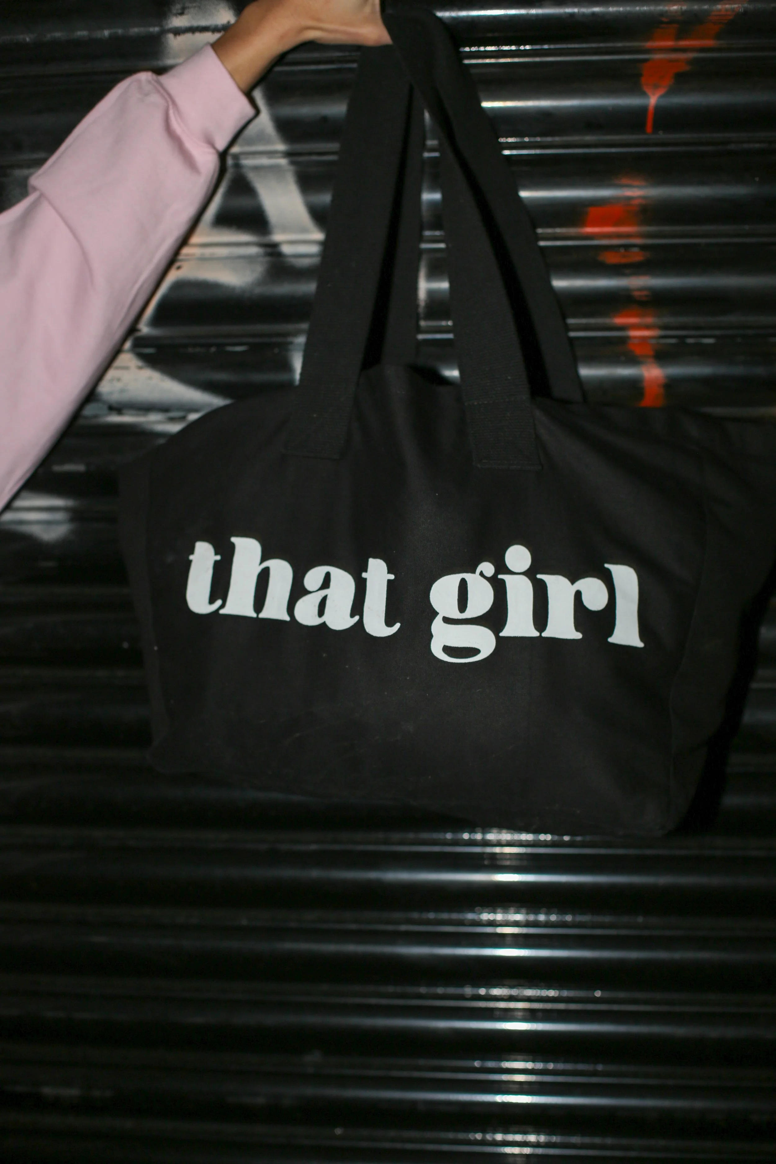 Black tote bag with white text that says 'that girl' being held up by a person's arm wearing a pink sleeve, against a black corrugated metal background.