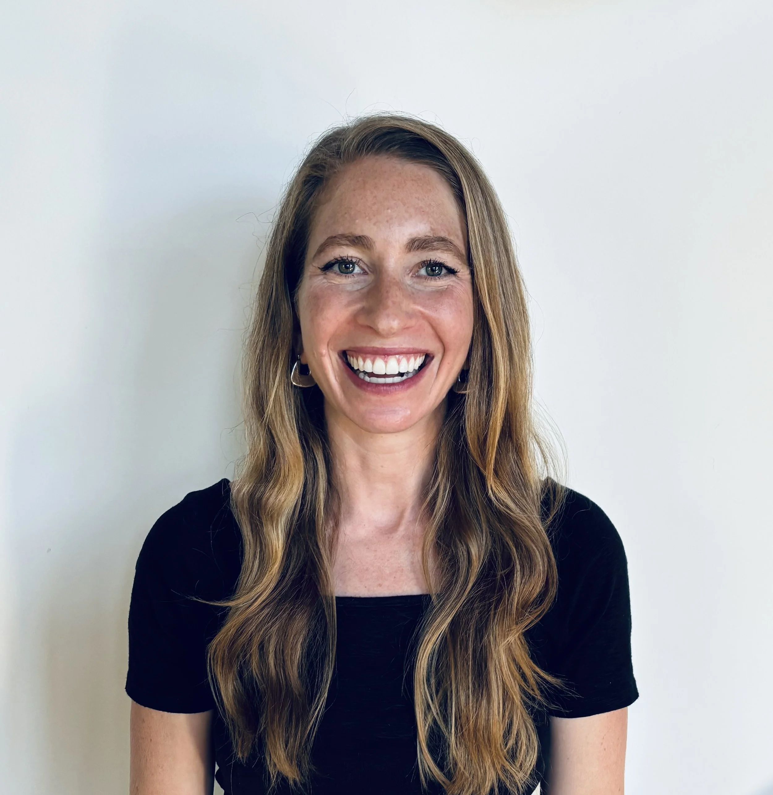 A woman in a wetsuit standing on a beach smiling, with surfers holding surfboards in the ocean behind her and cliffs in the distance.
