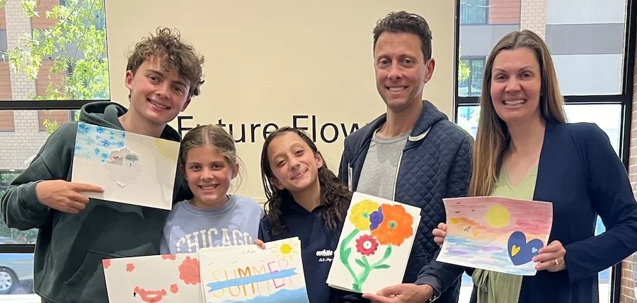 Group of five people, three children and two adults, holding painted pictures in an indoor setting with windows in the background.