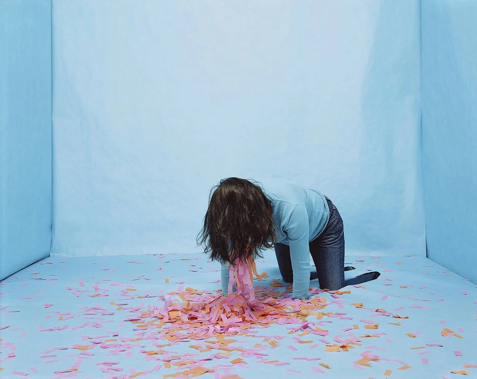 Person kneeling on the floor scooping up pink and orange confetti in a blue room with blue walls and blue floor.