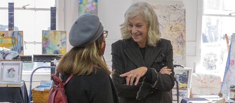 Two women talking and smiling in an art studio with colorful paintings in the background.
