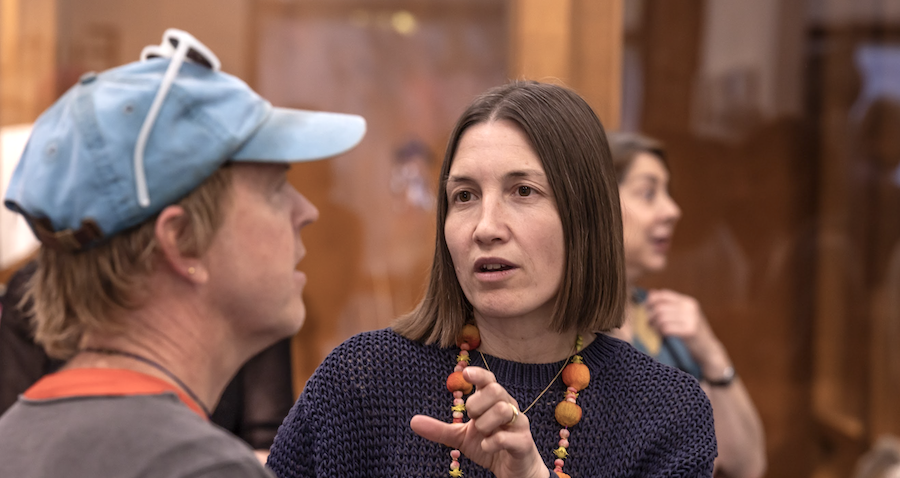 Two people conversing in a museum or gallery, with a woman in a blue sweater and an older woman with a light blue cap, and another woman in the background.
