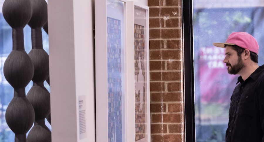 A man with a pink cap looks at colorful art or photographs displayed in a gallery or store window inside a brick-walled room, near a large window showing an outdoor scene.