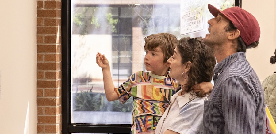 A family of three, including a young boy, a woman, and a man, are looking at a screen on a wall. The boy appears engaged, and the adults are smiling and interacting with him. They are indoors, near a window with an outdoor scene visible.