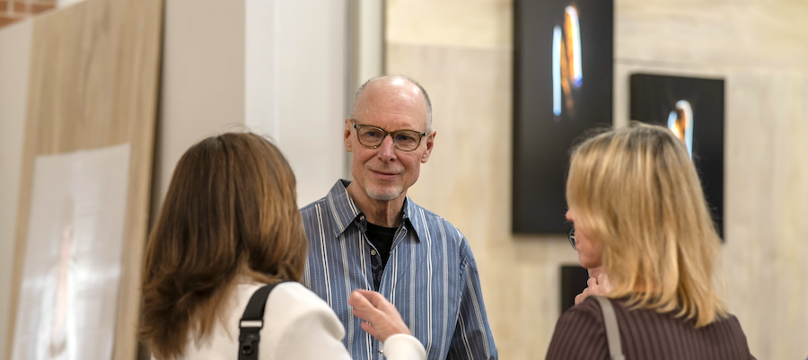 Three people engaged in conversation at an art gallery or exhibit space. Two women and a man with glasses are talking, with artwork visible on the wall behind them.