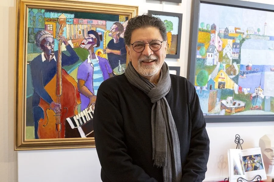 A smiling man with gray hair and glasses standing in an art gallery with colorful paintings behind him, including abstract cityscapes and a jazz band scene.