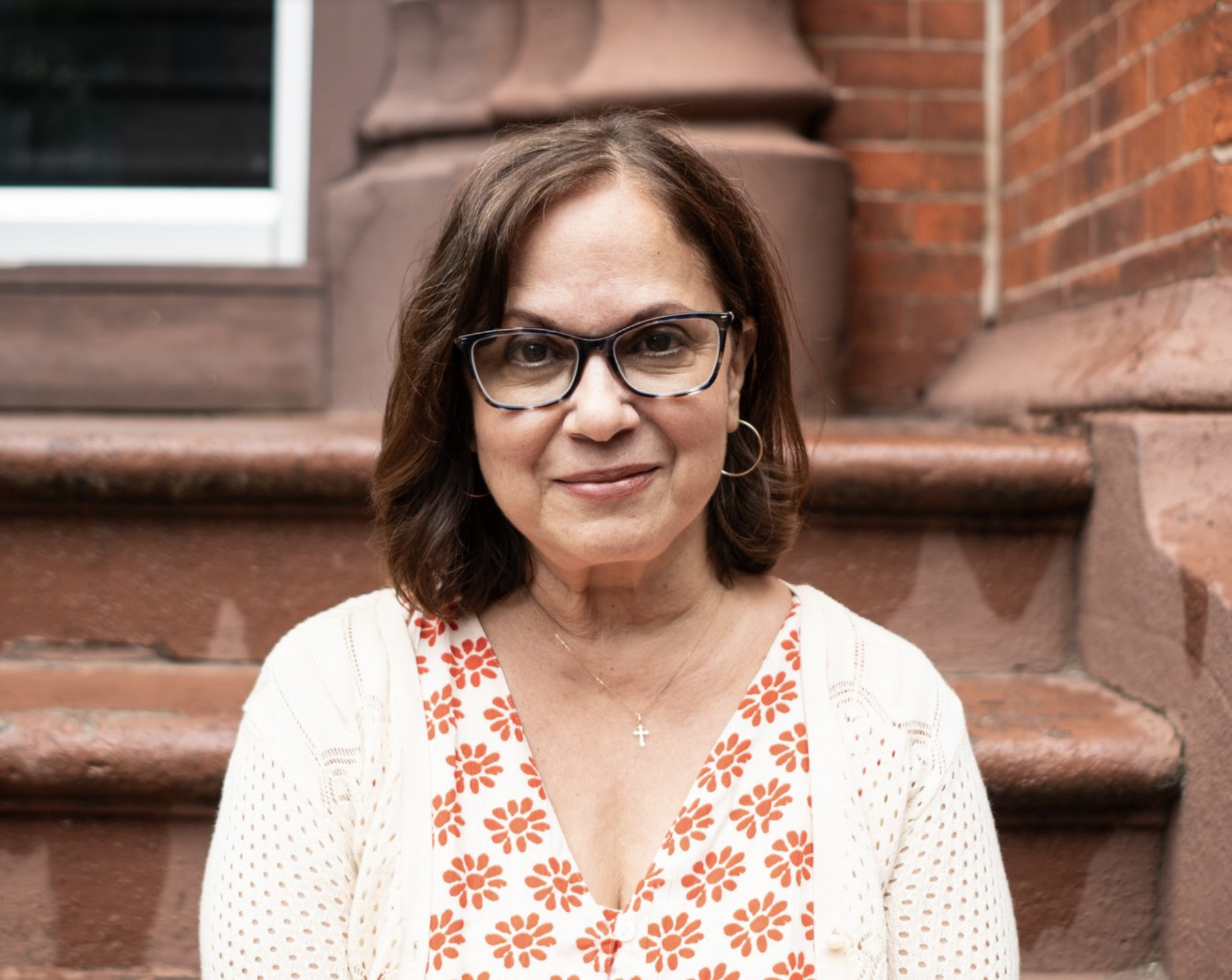 Woman sitting on steps outdoors, wearing glasses, a white sweater, and an orange floral top.