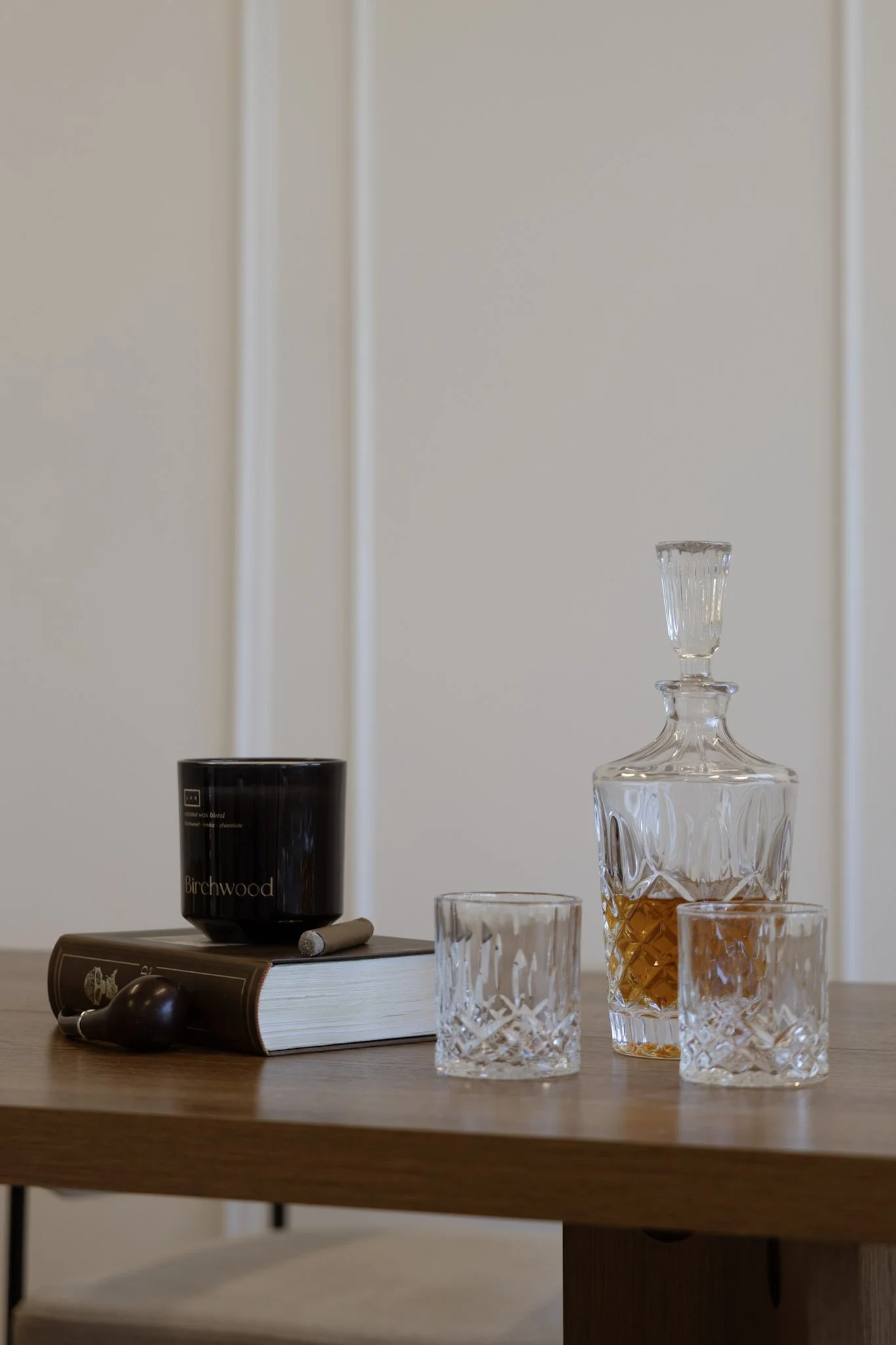 A wooden table with a black candle labeled Birchwood, a book, a silver spoon, a decanter with amber liquid, and two empty whiskey glasses.