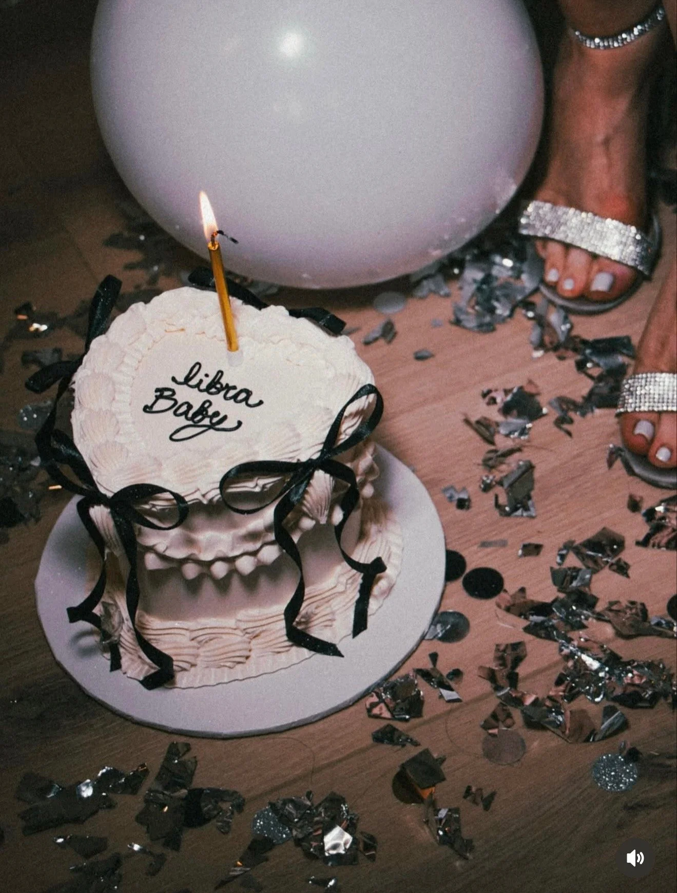A birthday cake with a candle and a message that reads 'Libra Babe' on top, surrounded by silver and black confetti on a wooden surface, with a large silver balloon and glittery sandals in the background.