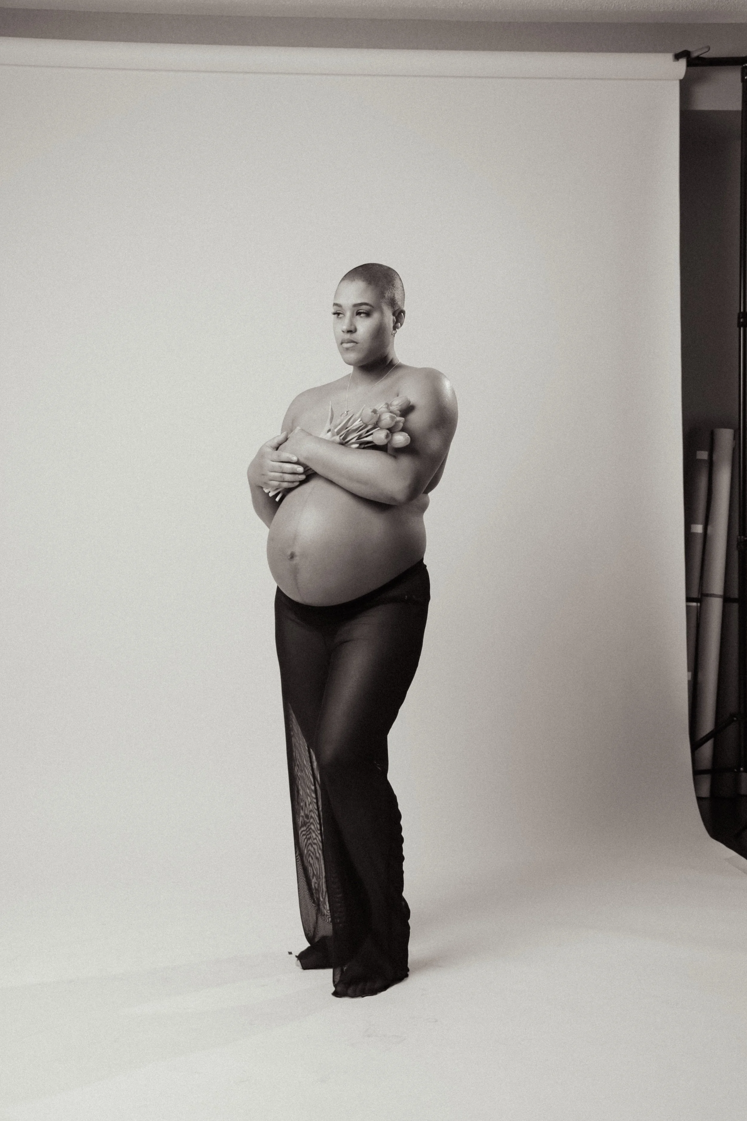 Black and white photograph of a pregnant woman standing in front of a plain background, holding a bouquet of tulips across her chest, wearing sheer black pants with a lace slit.