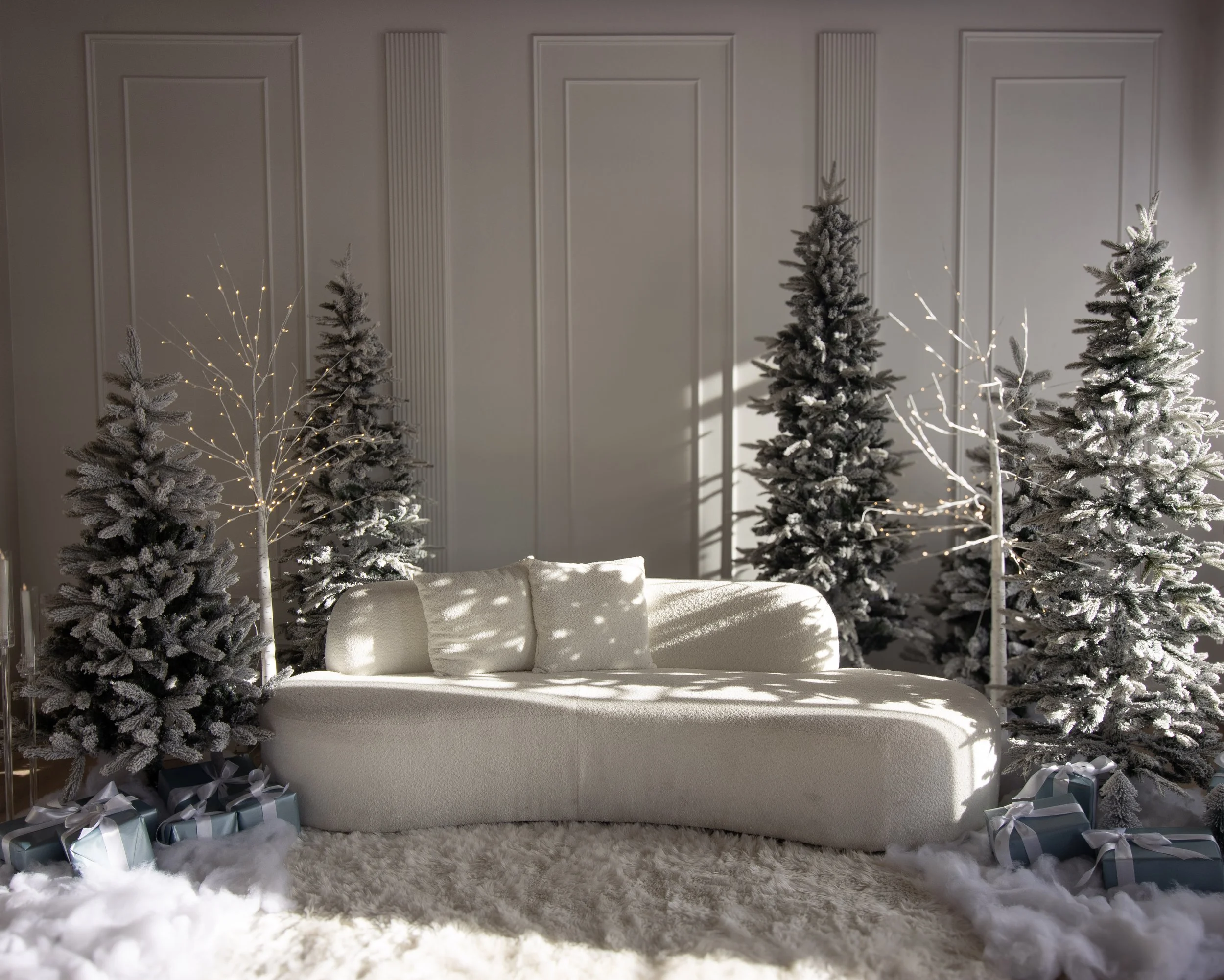 A cozy holiday scene featuring a white sofa with pillows, surrounded by snow-dusted Christmas trees with white lights, and wrapped presents on a white fluffy rug.