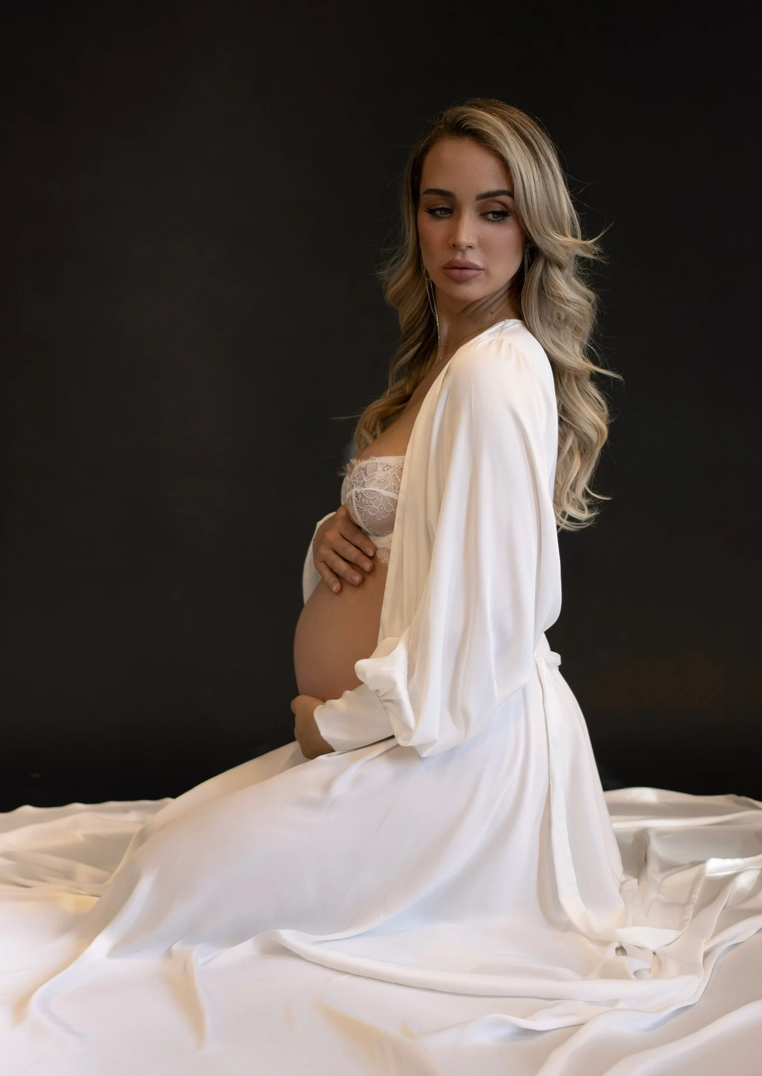 A pregnant woman with long wavy blonde hair, wearing a cream-colored satin robe, sitting on white fabric, against a dark background.