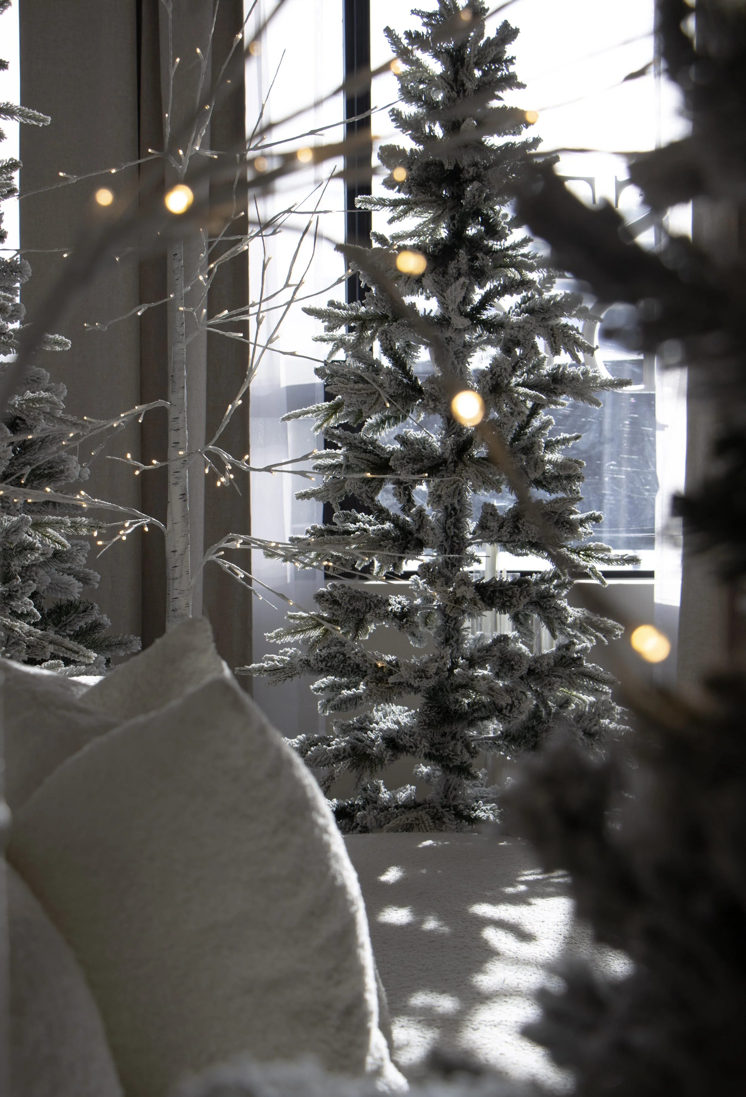 Cozy decorated Christmas scene with snow-dusted Christmas trees, pillows, and string lights inside a room with large windows.