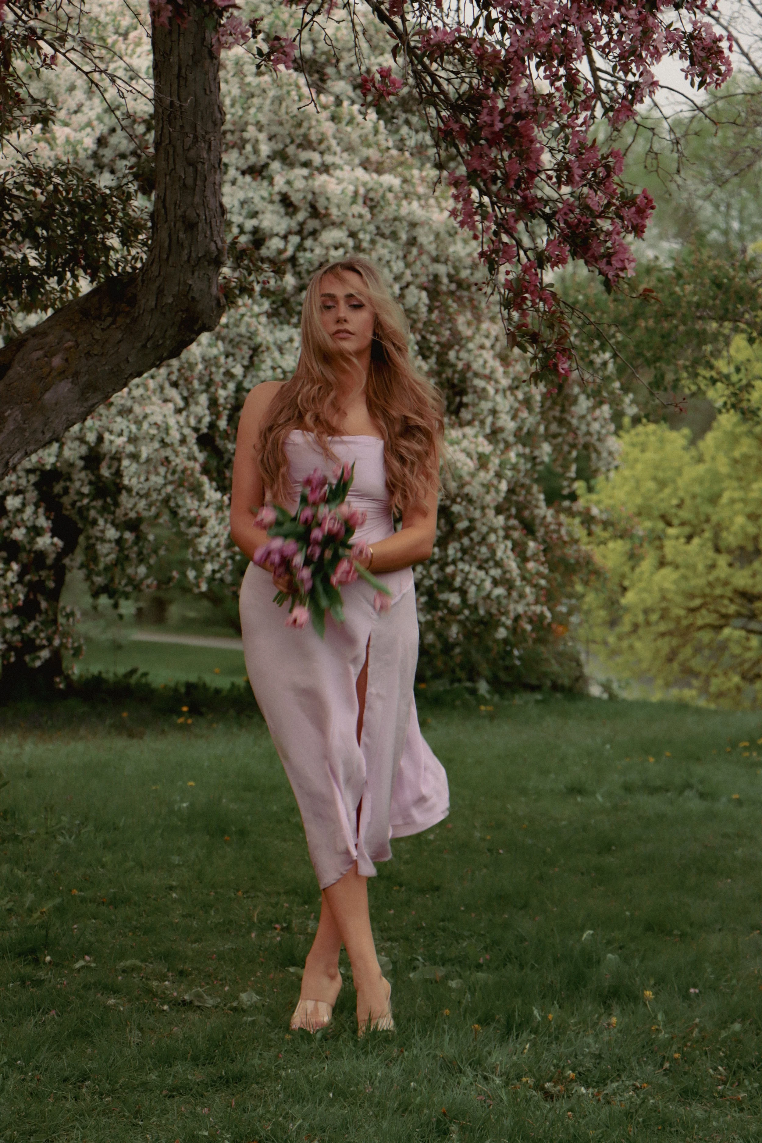 A woman with long wavy hair holding a bouquet of pink flowers, walking on grass in a garden with blooming trees and shrubs.