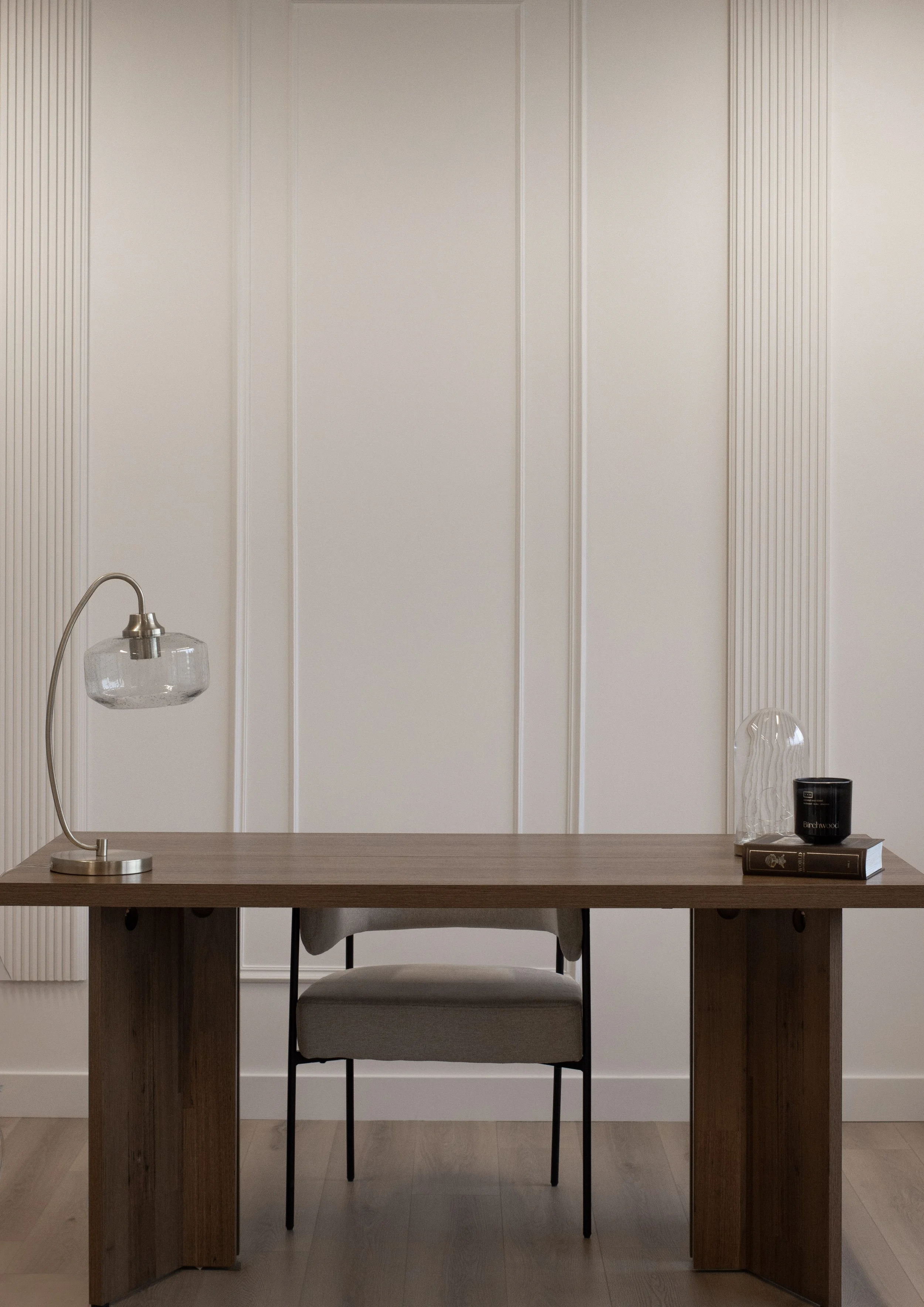 A minimalistic home office with a wooden desk, a beige chair, a modern glass desk lamp, and decorative items like a glass dome, candle, and books on the right side of the desk.