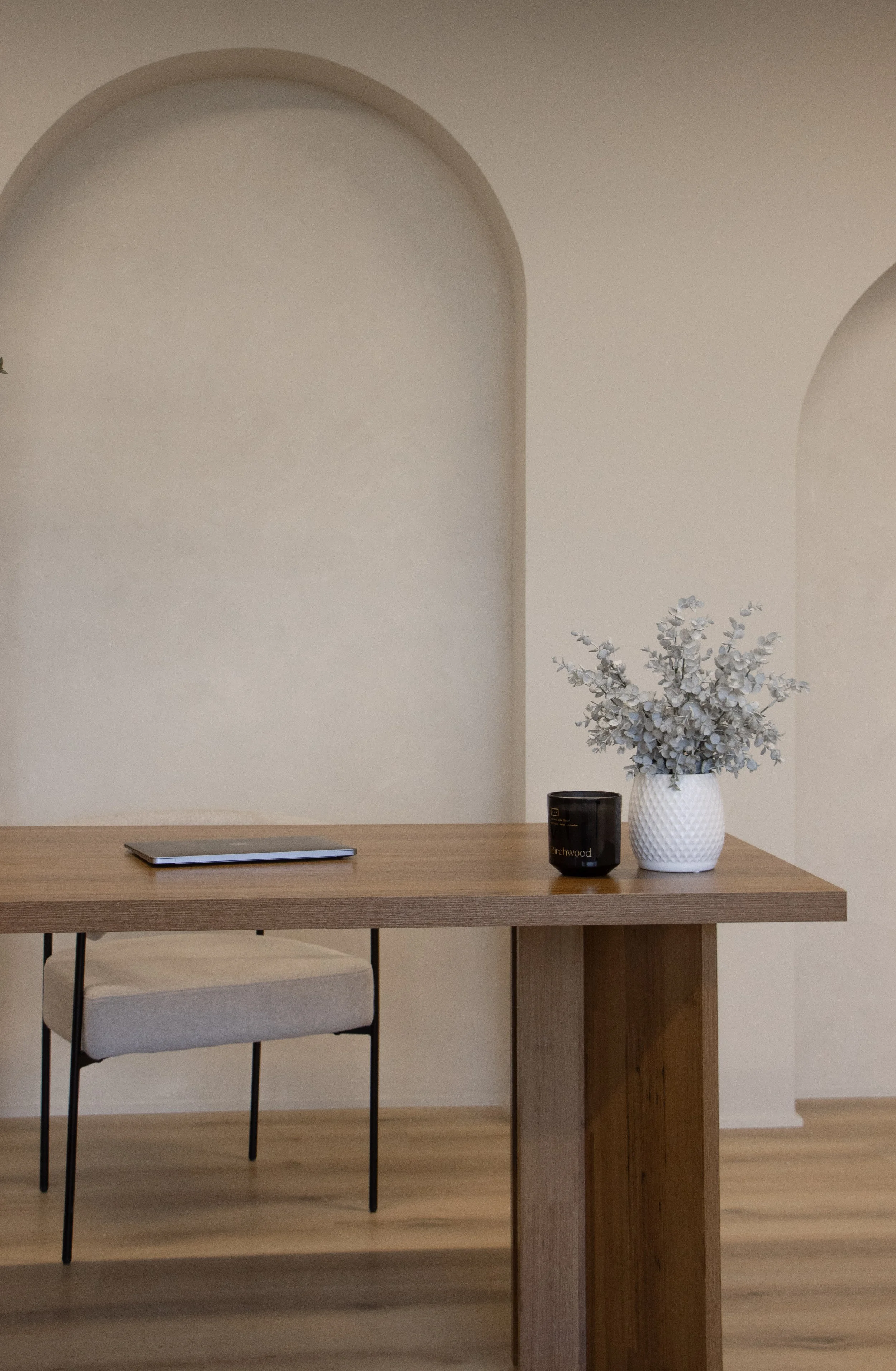A minimalist wooden desk with a beige chair behind it. On the desk, there is a closed laptop, a black candle, and a white textured vase with dried silver leaves. The background features light-colored walls with arched wall cutouts.