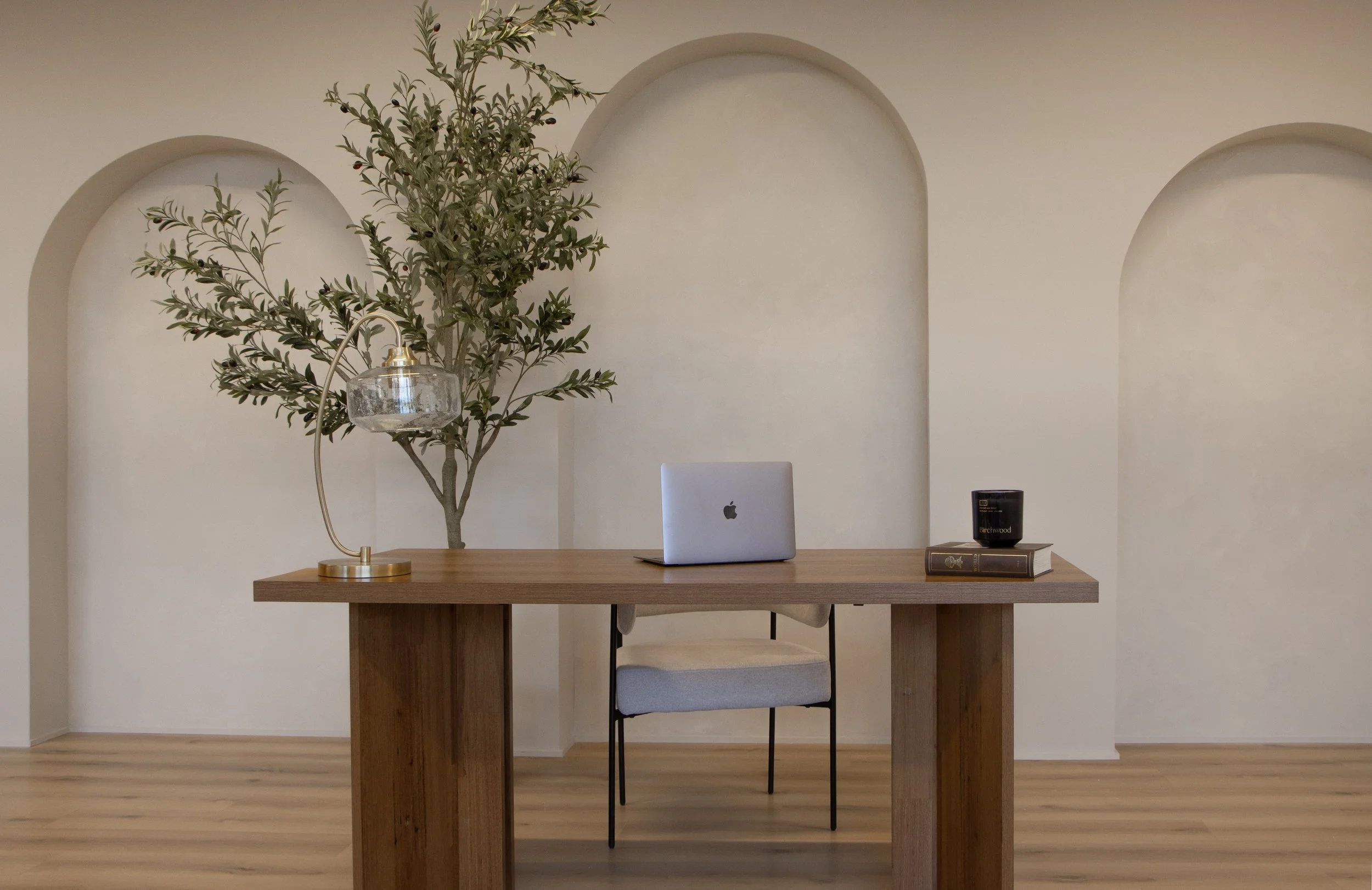Minimalist home office with a wooden desk, a closed silver MacBook, a black candle holder on top of a book, a beige chair, a decorative lamp, and a tall houseplant against a cream wall with arches.