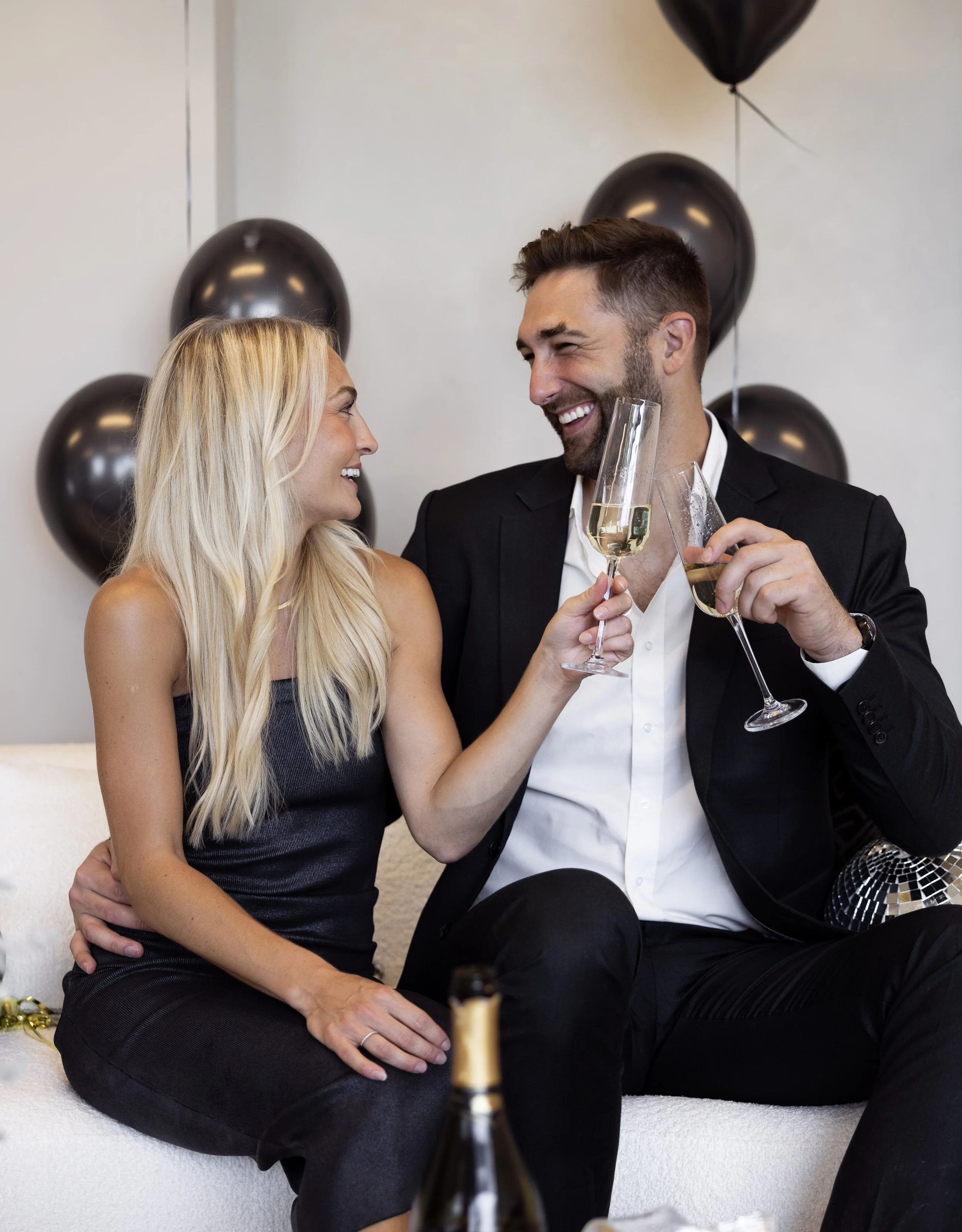 A man and woman sitting close together, smiling at each other and toasting glasses of champagne at a celebration party with black balloons behind them.
