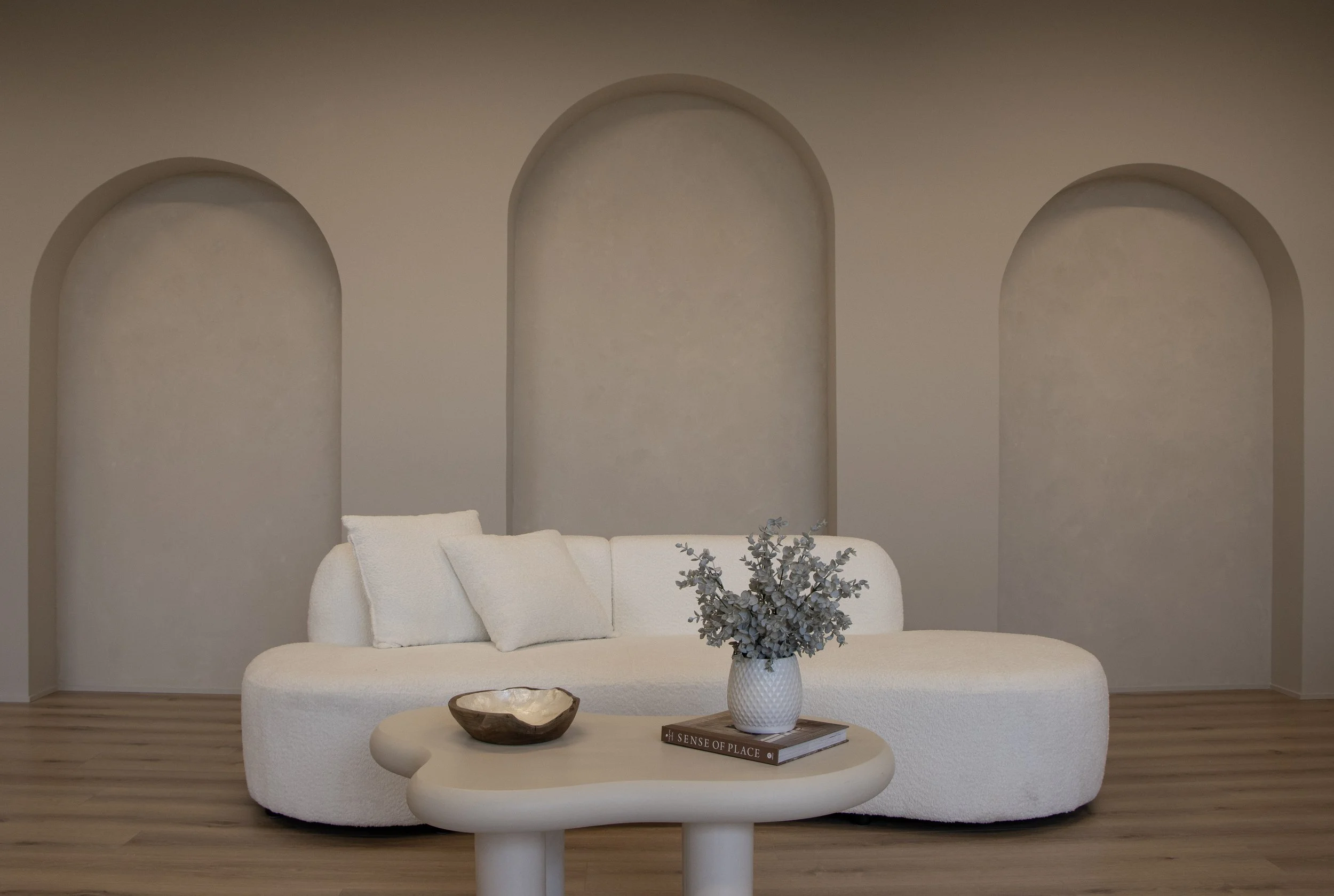 Minimalist living room with a white curved sofa, a cream-colored coffee table with a wooden bowl and a white vase of eucalyptus, and three arched wall niches in a neutral color.