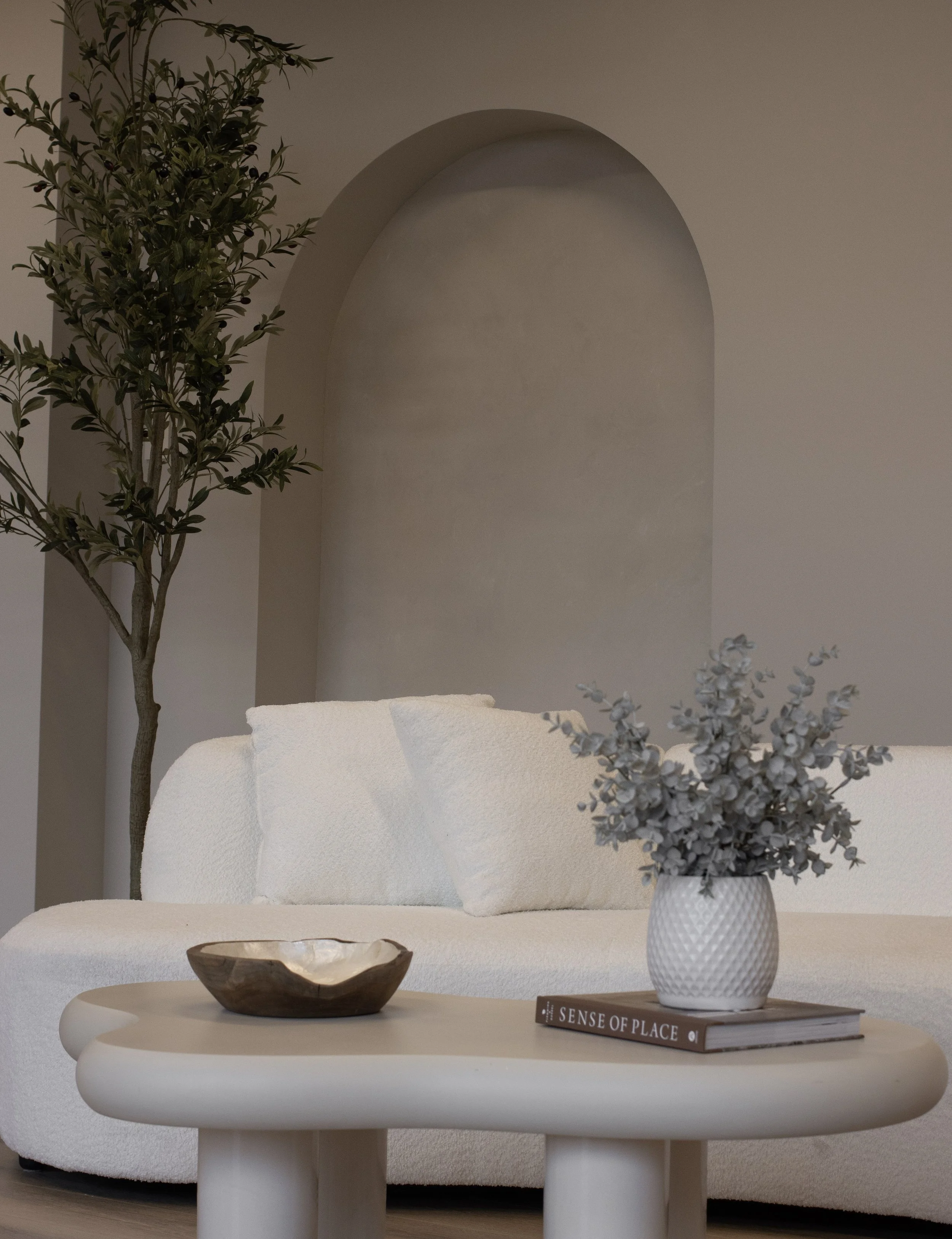 A cozy living room corner with a white textured sofa, a round white coffee table holding a decorative wooden bowl and a white vase with grey leaves, a book titled 'Sense of Place,' and a tall green plant in the background near a wall with an arched r