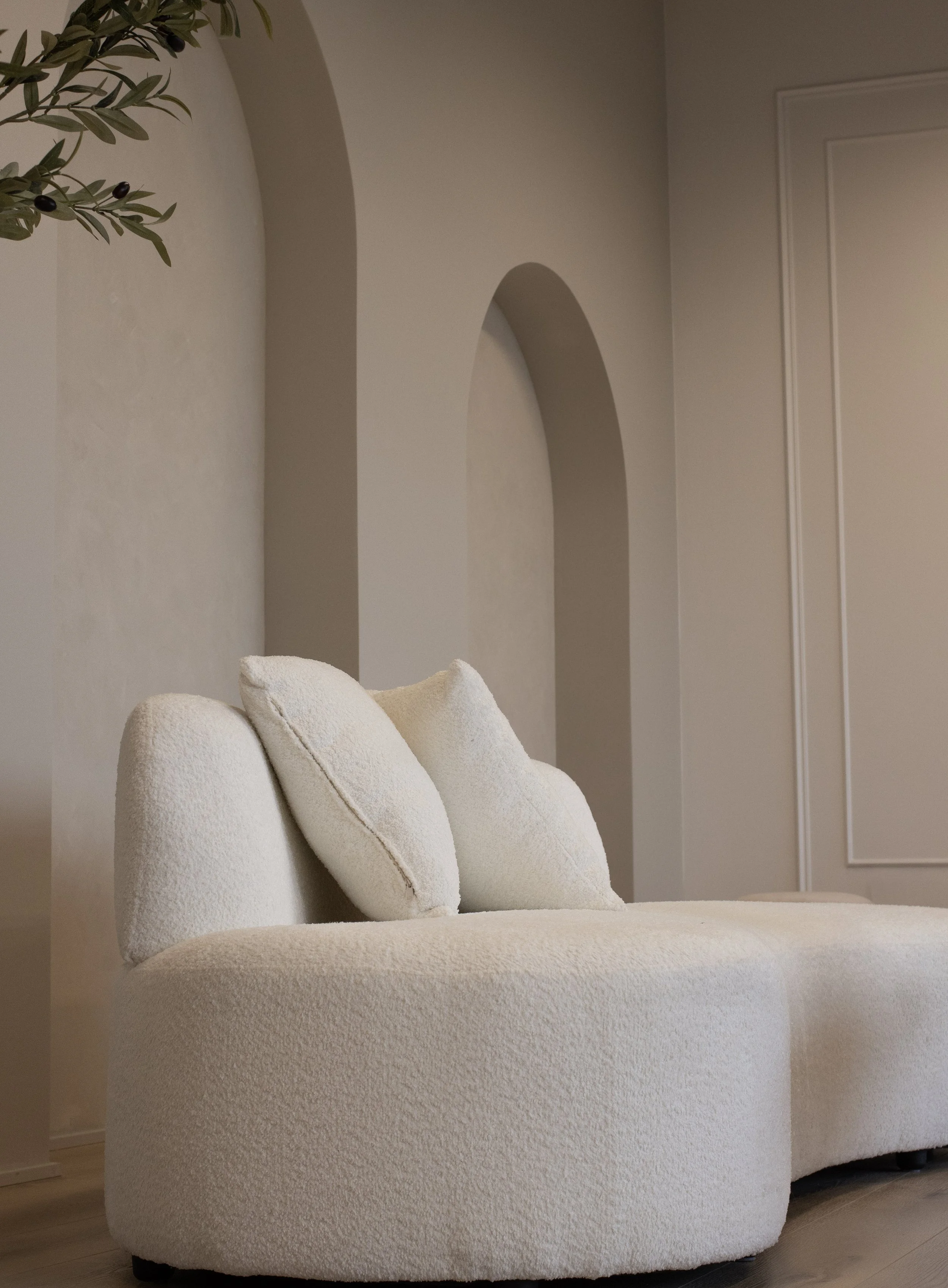 Living room corner with a white textured sofa and two matching pillows, beige wall with arch and rectangular wall panel.