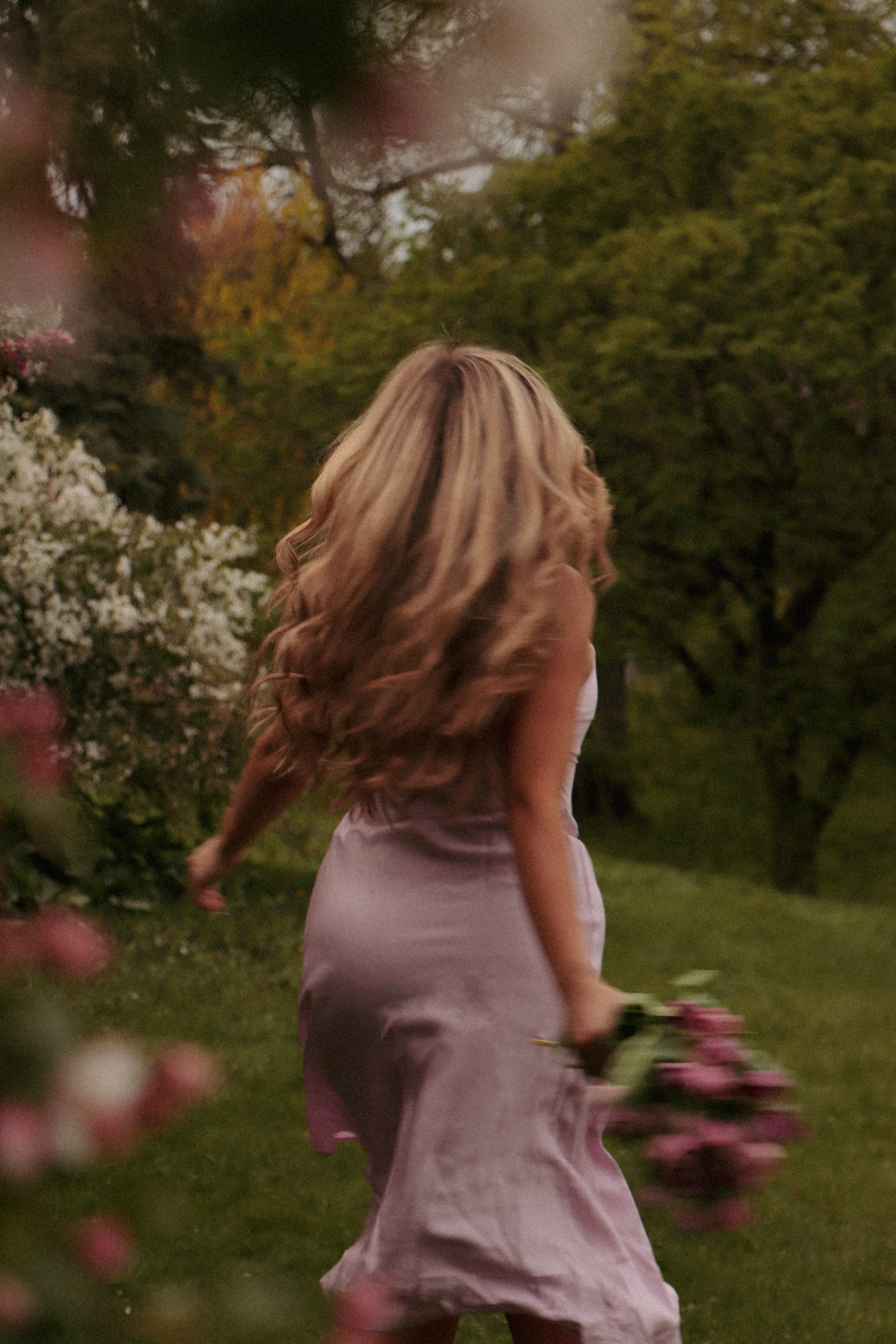 A woman with long, wavy blonde hair is walking through a garden, holding a bouquet of pink flowers, with trees and blooming bushes in the background during dusk.