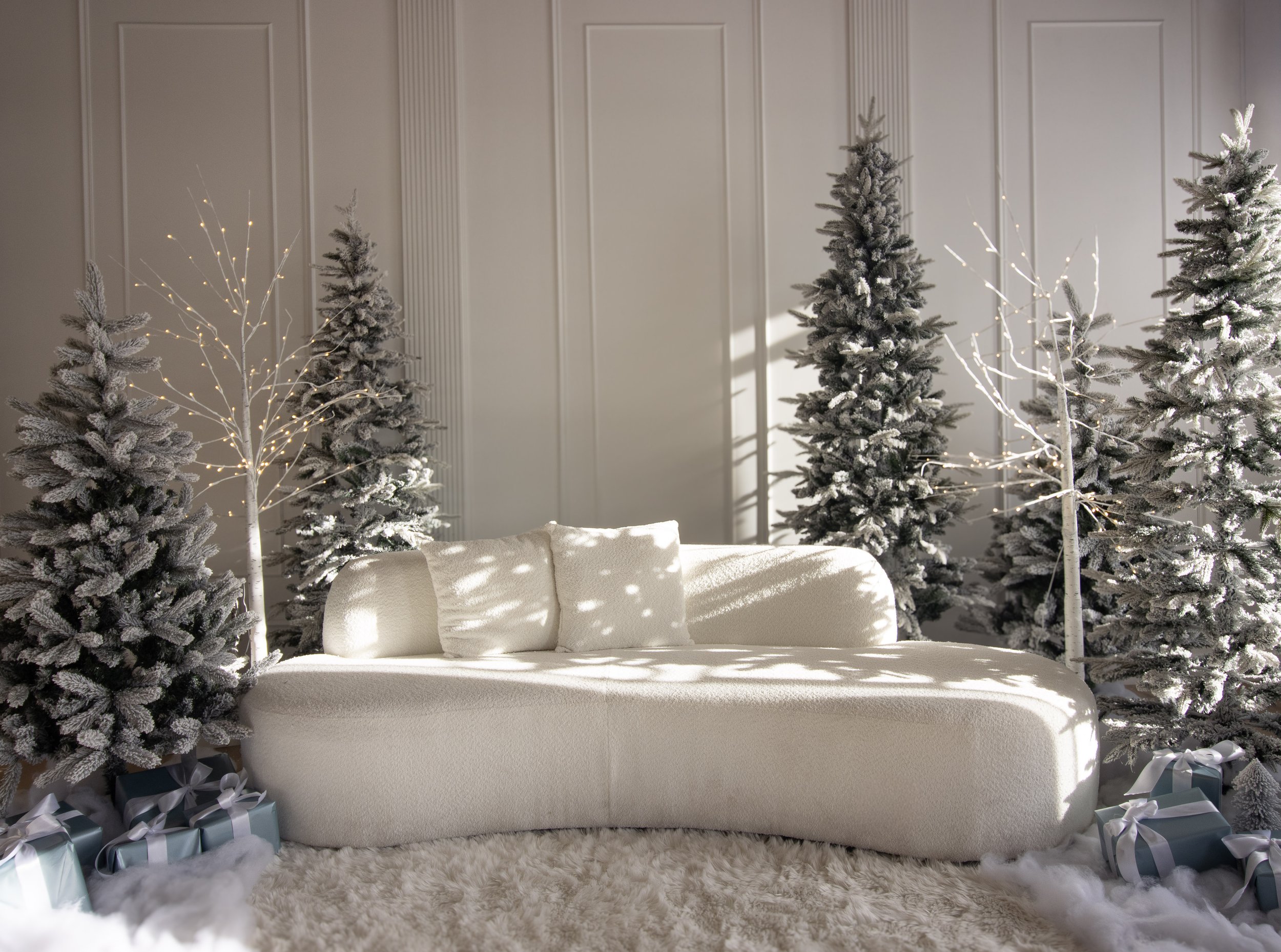 A cozy holiday scene with a white sofa in front of Christmas trees decorated with snow-like flocking, surrounded by wrapped gifts on a plush rug.