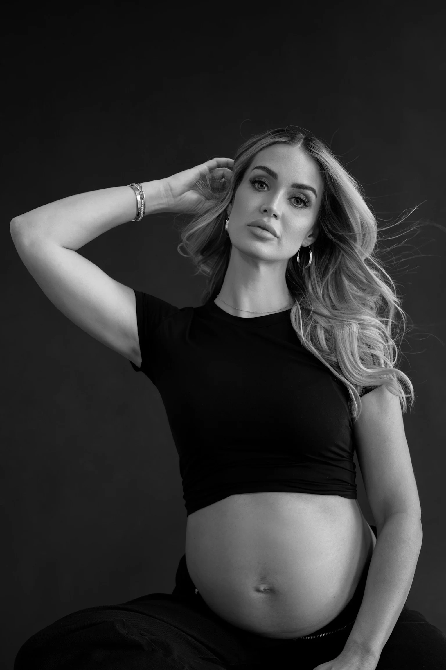 A pregnant woman with long, wavy hair, wearing hoop earrings, bracelets, and a black crop top, sitting against a dark background.