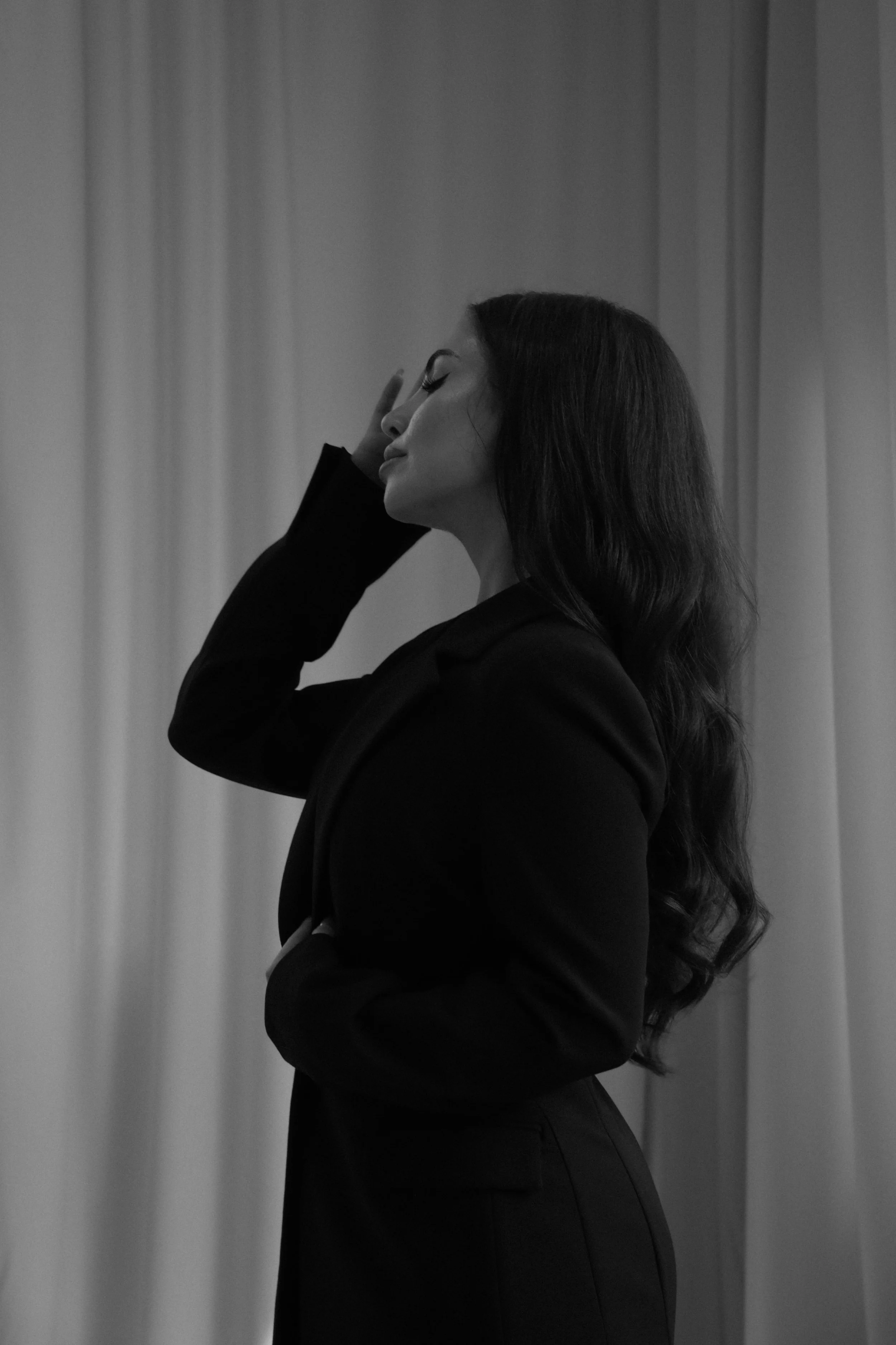 A black-and-white photo of a woman with long wavy hair, wearing a dark blazer, standing with her eyes closed and touching her face with her right hand, against a plain curtain background.