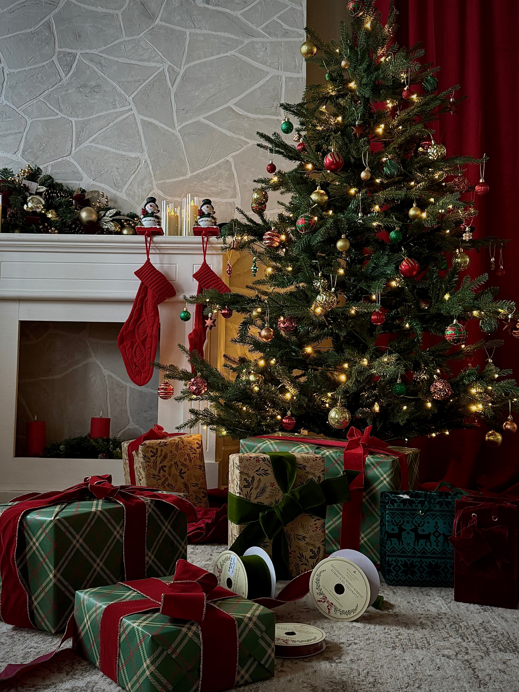 A decorated Christmas tree with gold, red, and green ornaments and lights, surrounded by wrapped gifts with ribbons, in a cozy living room with a stone fireplace, Christmas stockings, and candles.