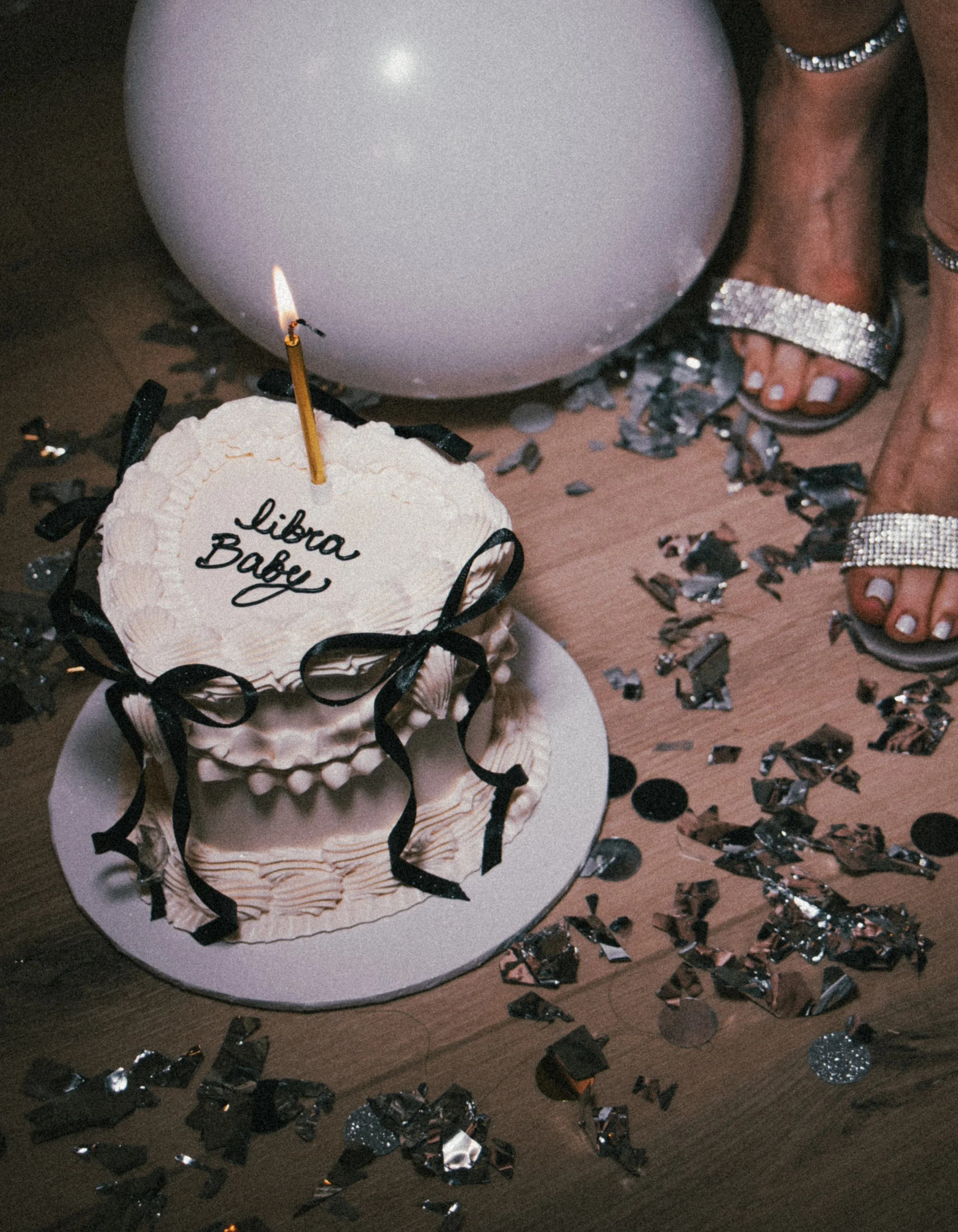 Celebratory scene with a small birthday cake labeled 'Libra Baby' topped with a single lit candle, surrounded by silver and black confetti, and a large white balloon. Part of a woman's foot in silver heels is visible in the background.