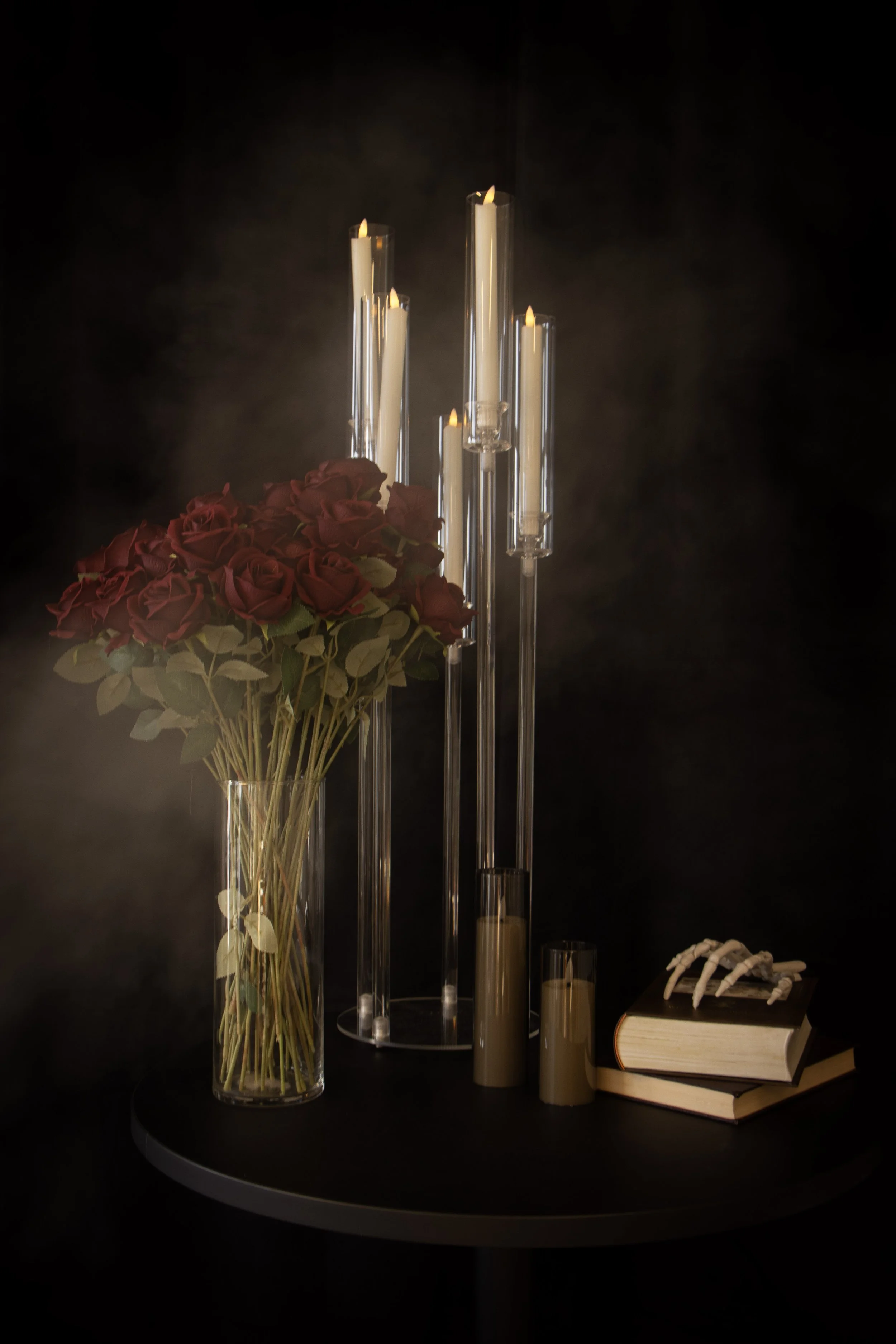 A black table with a large bouquet of dark red roses in a glass vase, several tall candles in glass holders with flames, and stacked books with a decorative bone or claw on top, against a dark background.