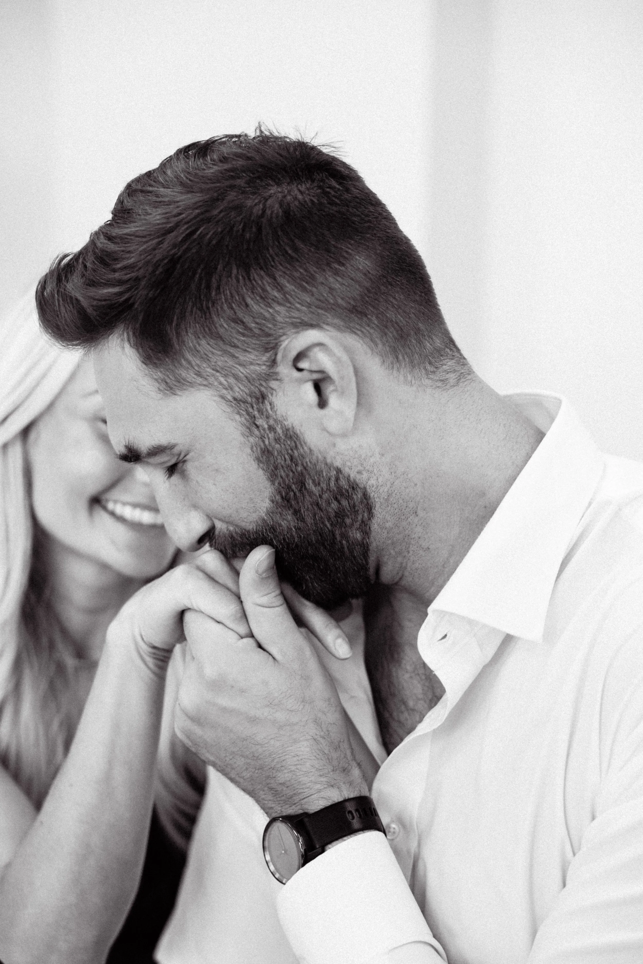 A close-up black-and-white photo of a man with a beard and short hair holding hands with a woman, both smiling softly, with their foreheads touching.