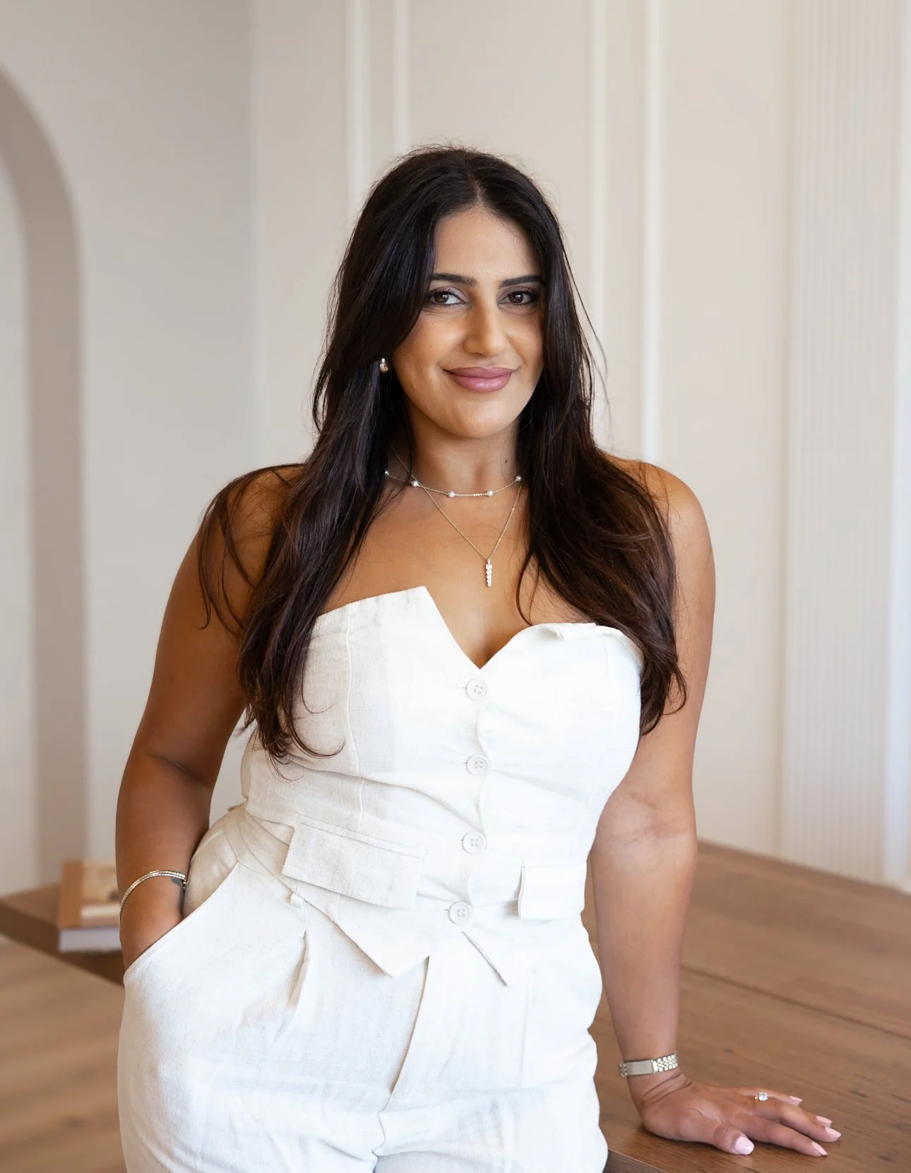 A woman with long dark hair, wearing a white sleeveless button-up top and matching pants, standing in a room with neutral-colored walls.