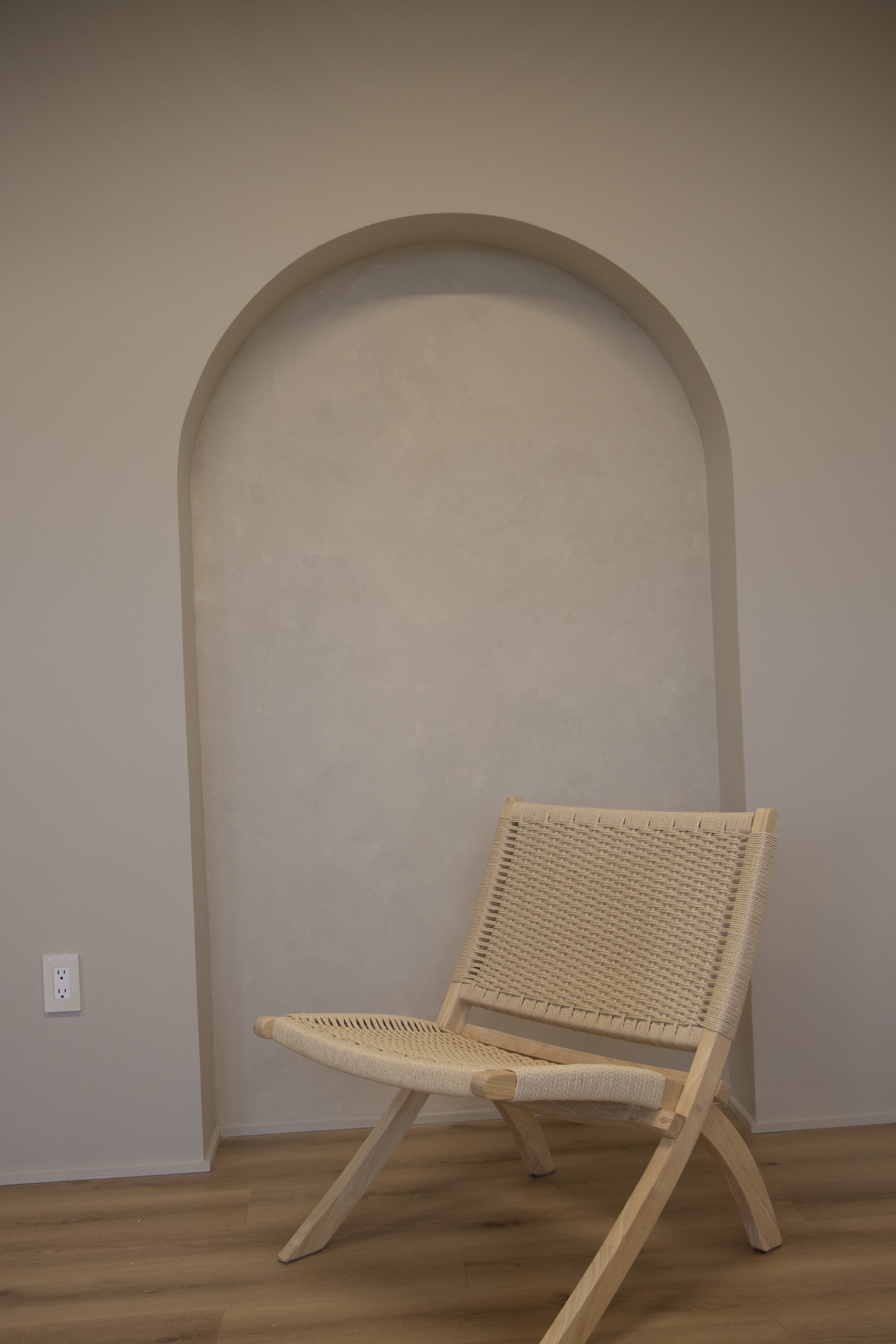 A beige woven chair with wooden legs in front of a beige arched niche in a room with wooden flooring.
