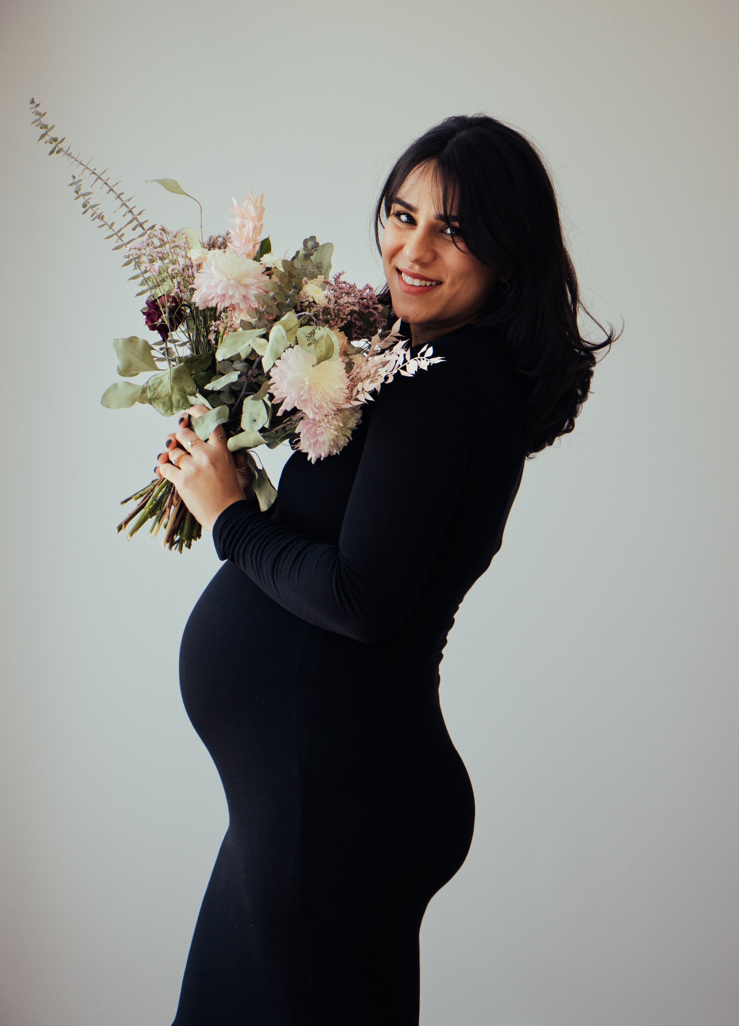 A pregnant woman with dark hair, wearing a black outfit, smiling, and holding a bouquet of flowers, standing against a plain light-colored background.